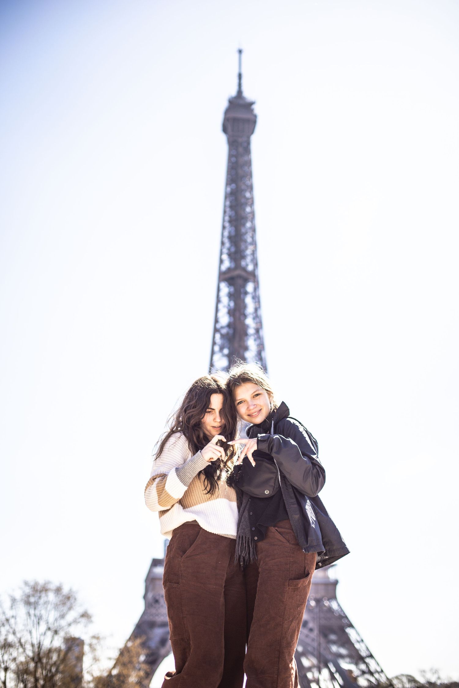 Two women embracing and smiling with the Eiffel Tower visible against a bright sky in Paris.