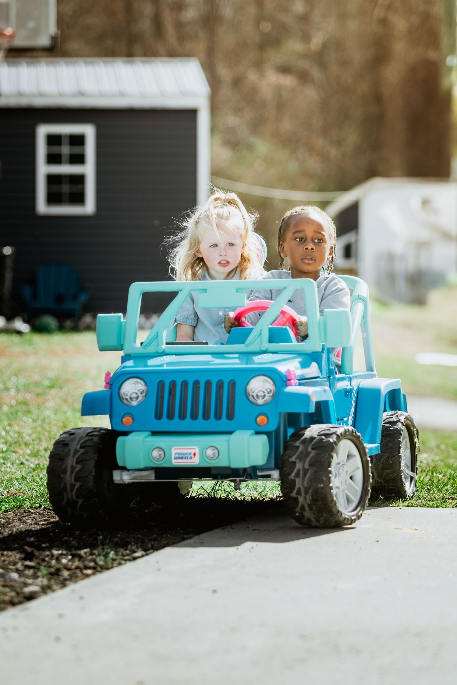 Two young girls ride a blue Power Wheels toy Jeep on a paved driveway outdoors.