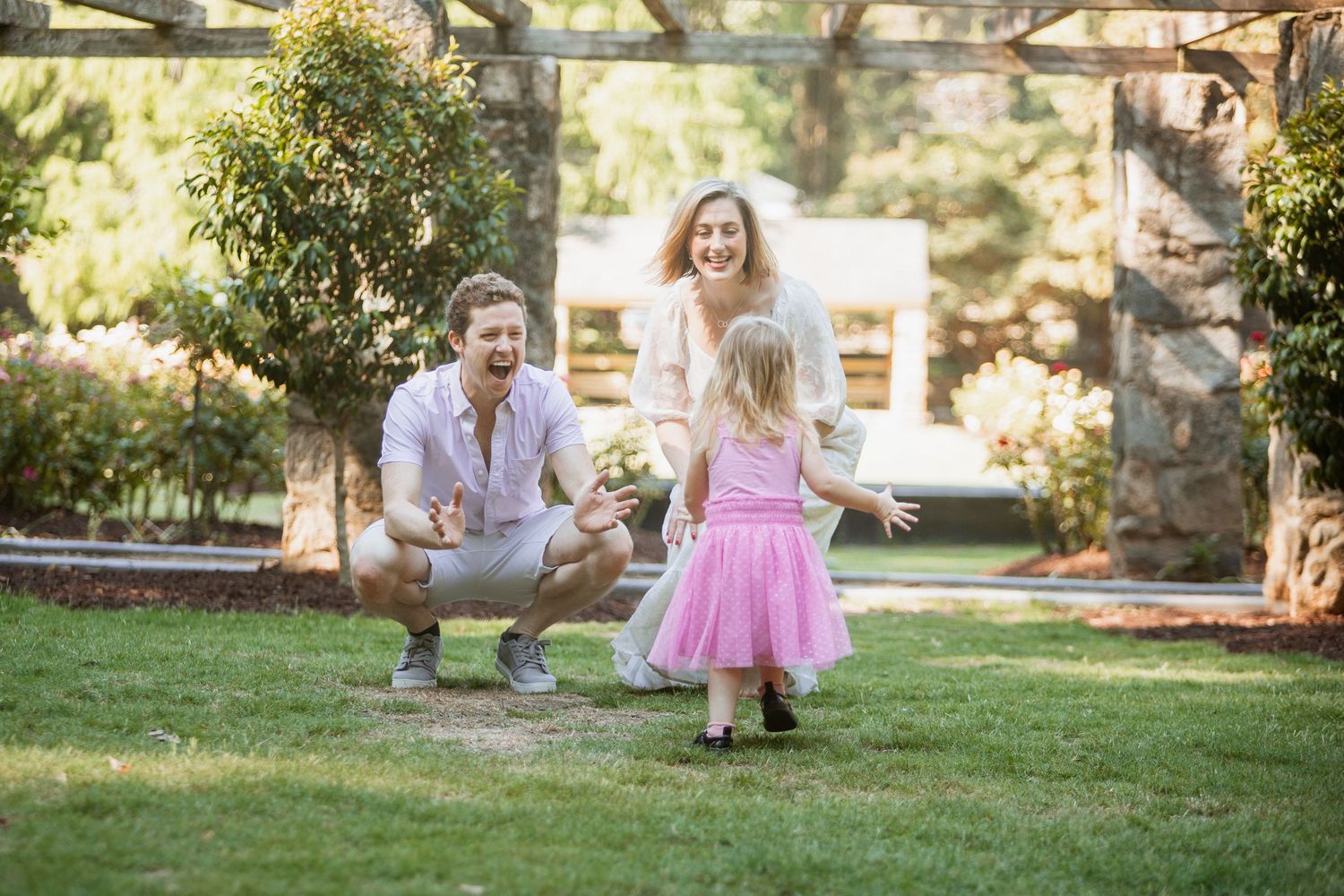 Young girl in pink dress running toward crouching father as mother laughs in garden.