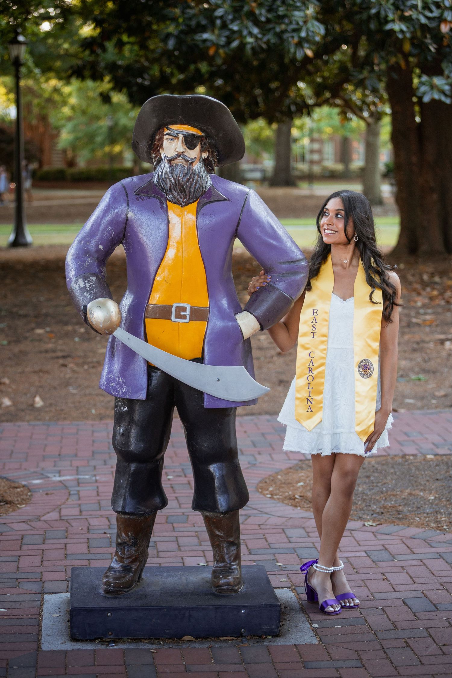ECU graduate in white dress smiles beside the purple and gold Pirate mascot statue on campus.