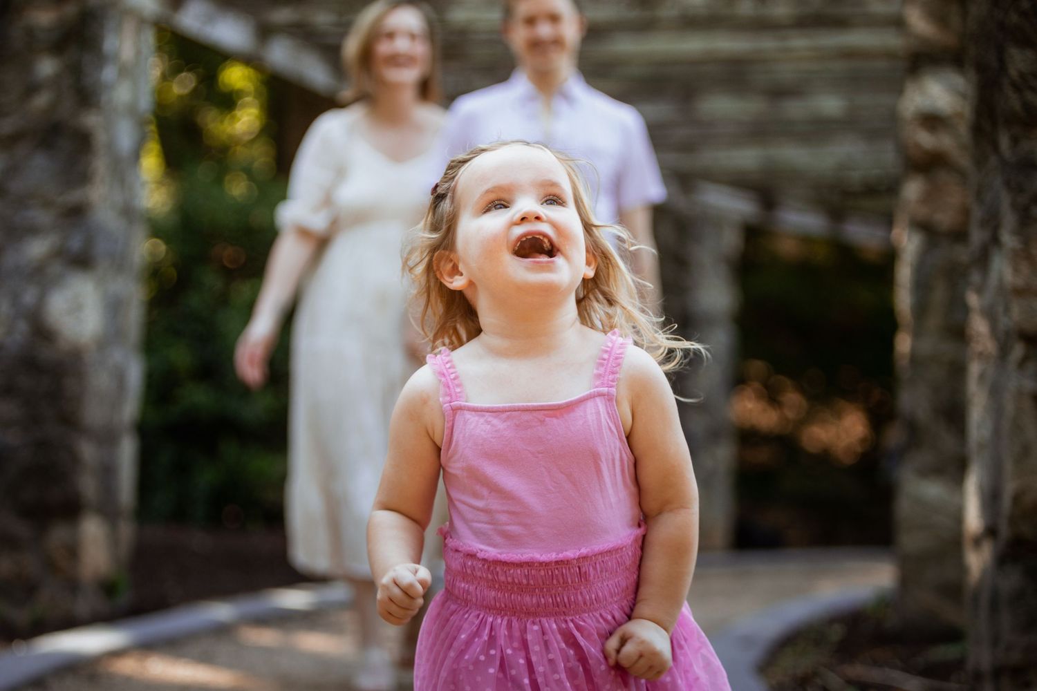 Joyful toddler girl in pink dress laughing outdoors with parents walking in background near log cabin.