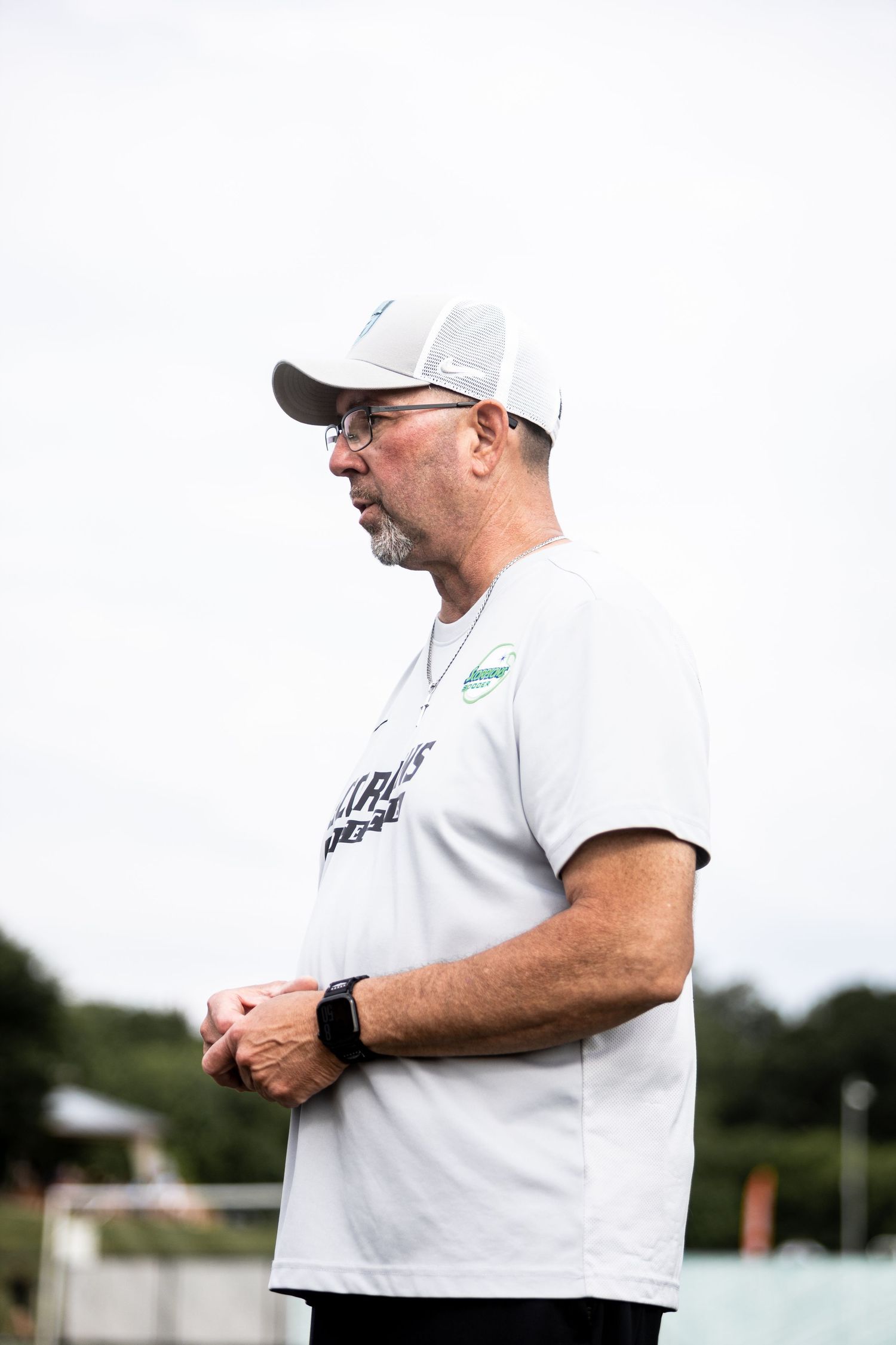 Male soccer coach in white cap and shirt observes practice from sideline with hands clasped.