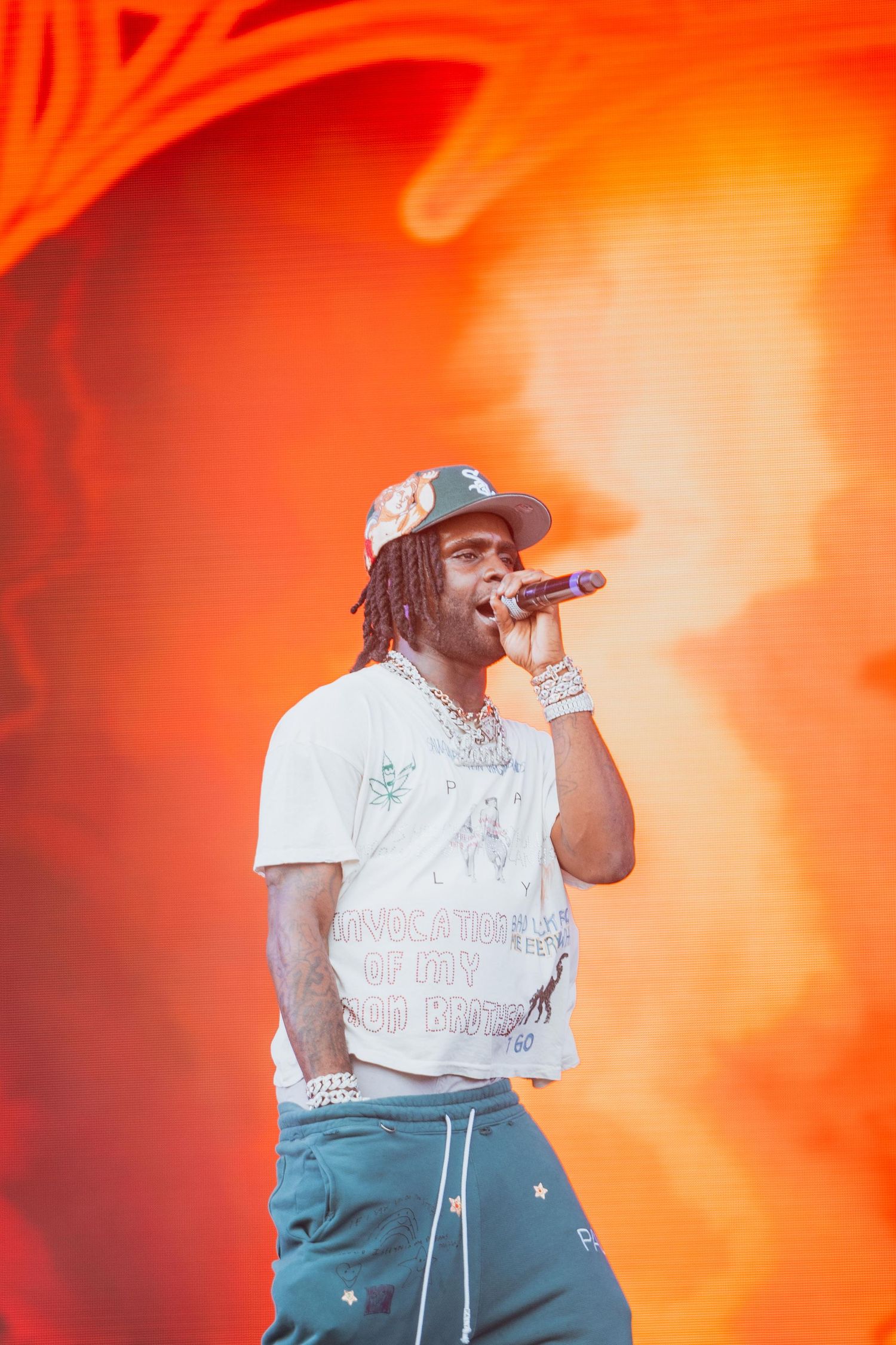 Rapper singing into microphone on stage wearing white graphic tee, teal star-print pants, and chains.