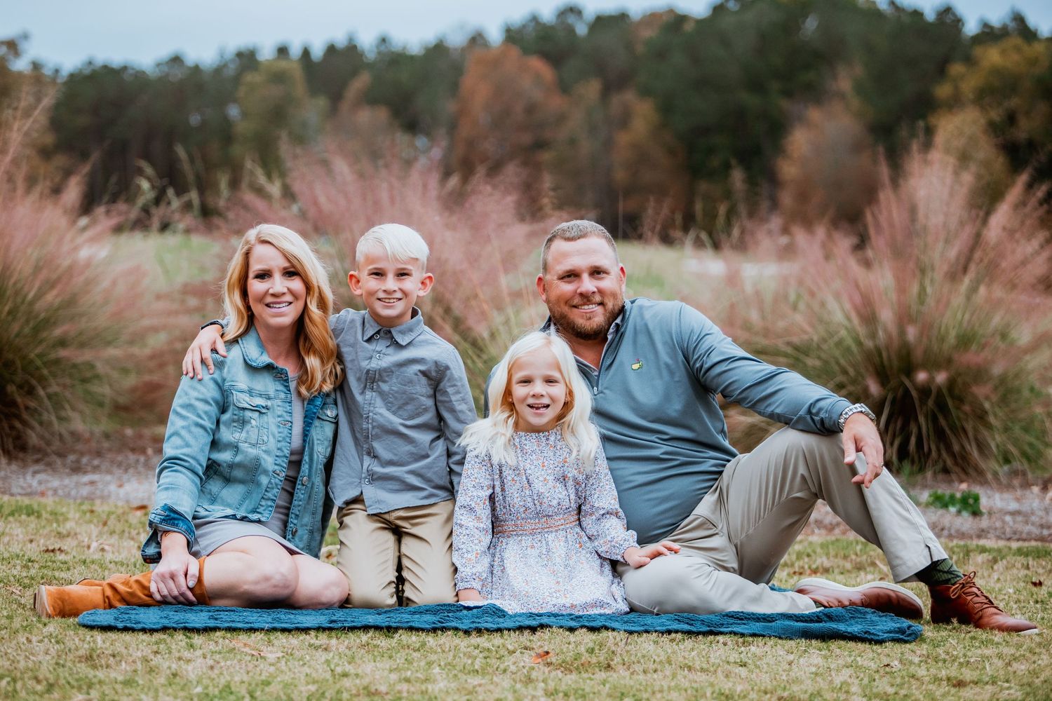 Cheerful family of four sits together on blue blanket in a field with colorful fall grasses behind.