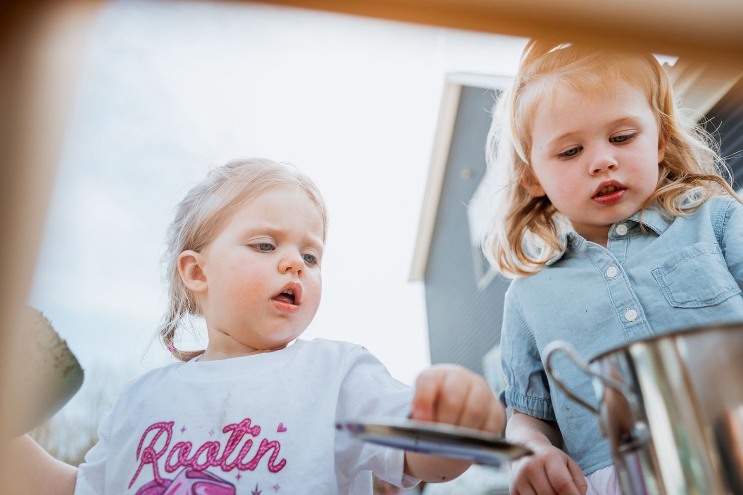 Two young blonde girls playing with a metal pot outdoors on a sunny day.