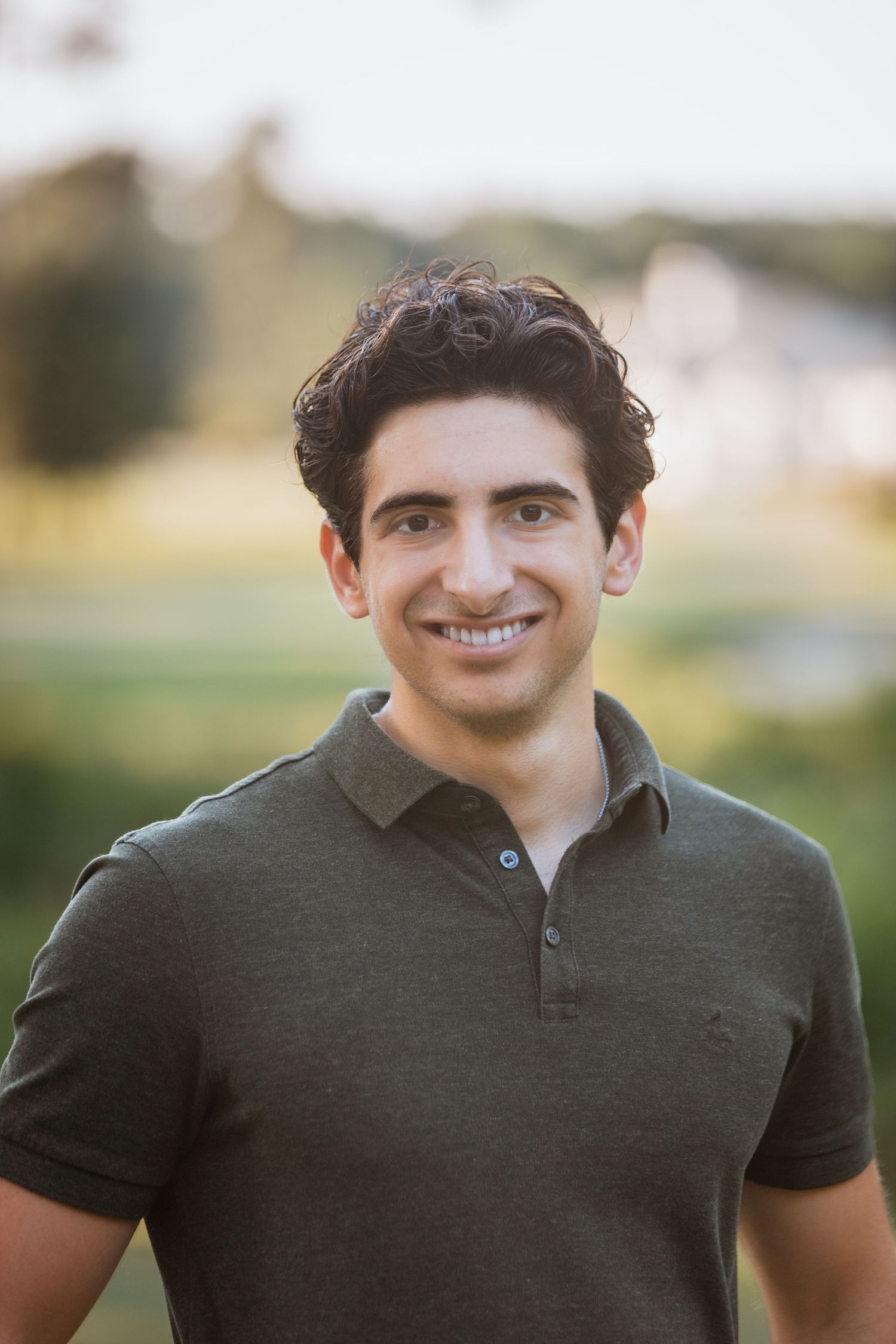 Young man with dark curly hair smiling outdoors in olive green polo shirt.