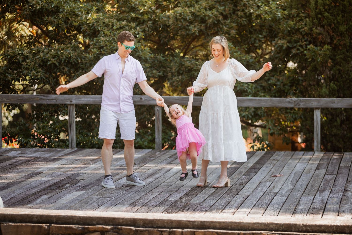 Parents holding hands with young daughter in pink dress on wooden deck outdoors.