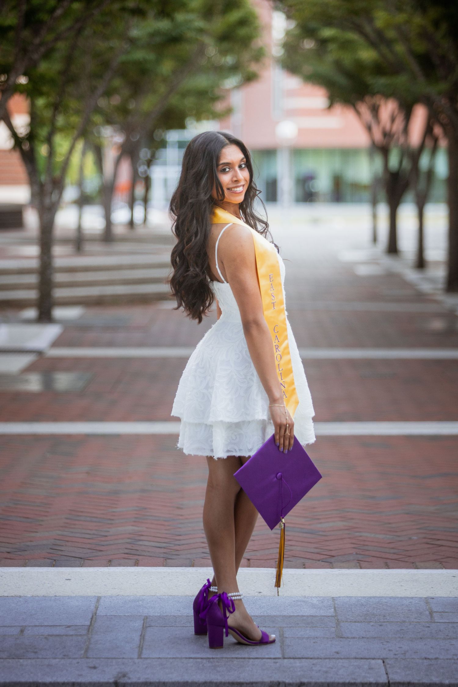 Smiling graduate in white dress holds purple cap and gold stole on ECU campus walkway.