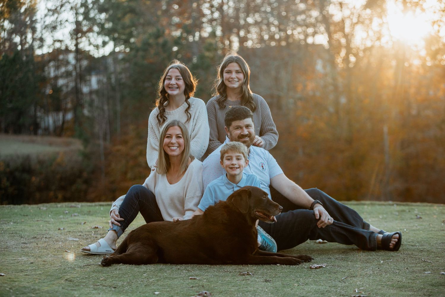 Smiling family posing with their chocolate Lab dog in a sunlit outdoor autumn setting.