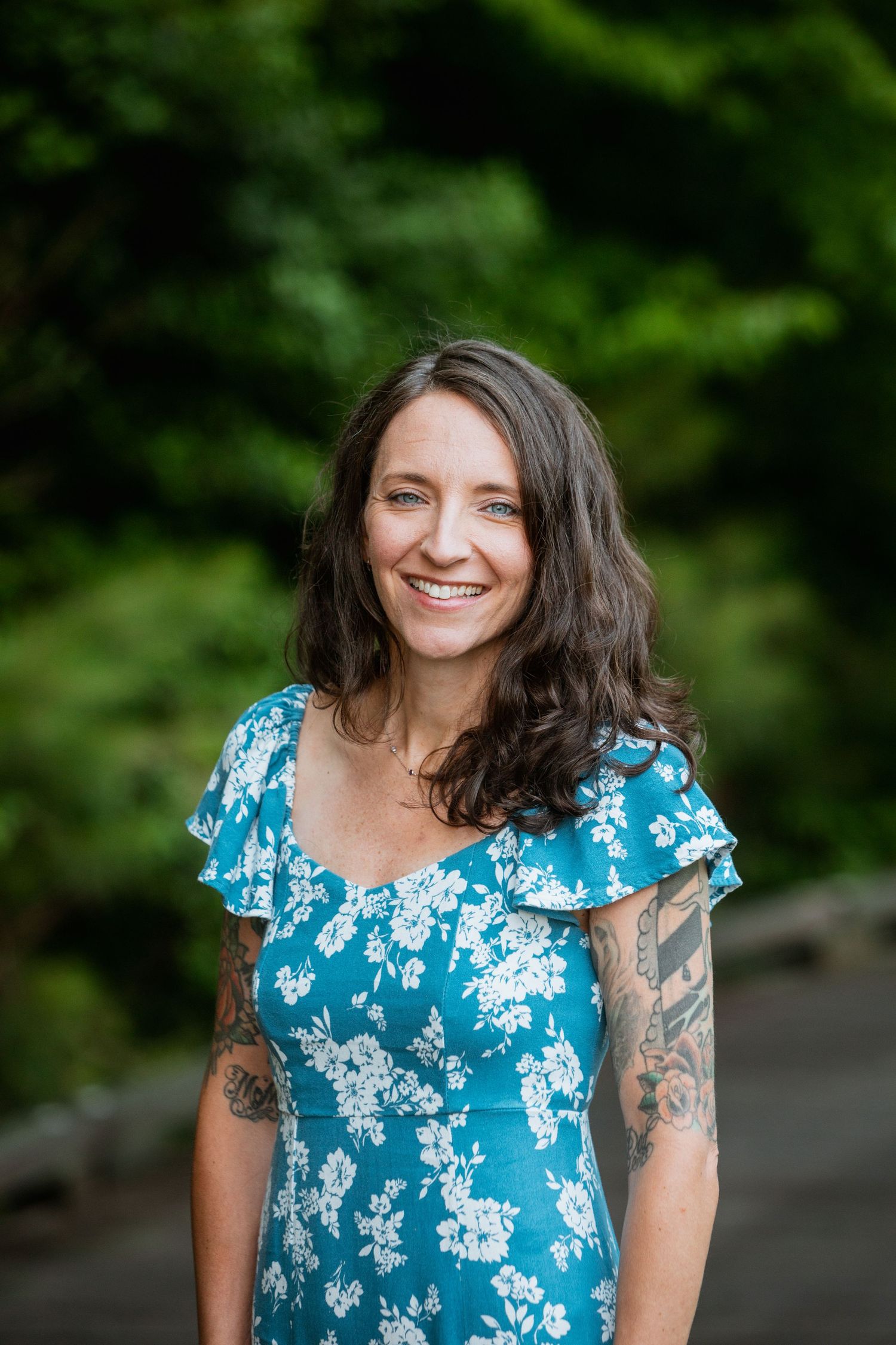 Smiling brunette woman with tattoos wearing a blue floral dress posing outdoors with green trees behind her.