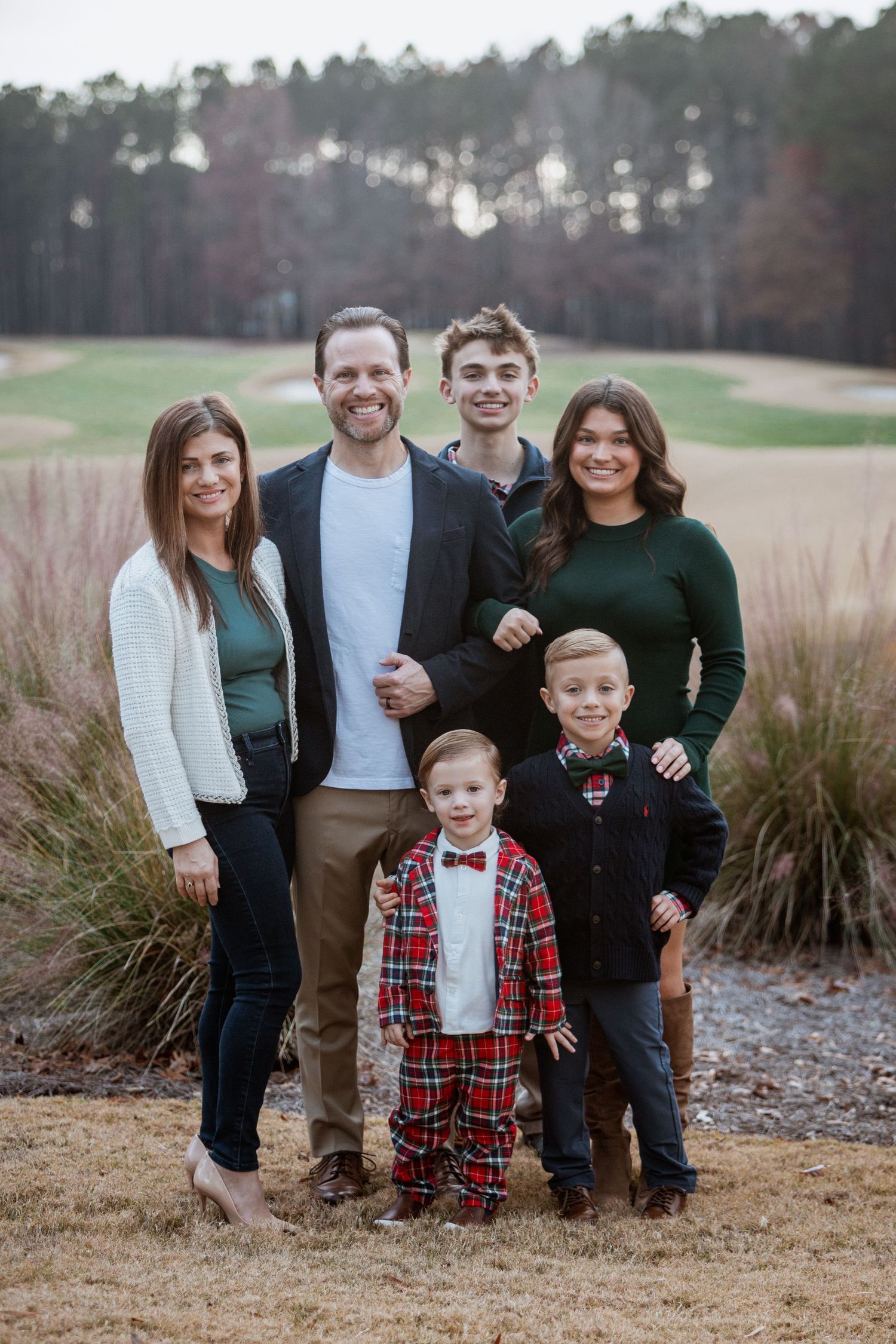 A family of six smiles together outdoors near a golf course on a cool winter day.