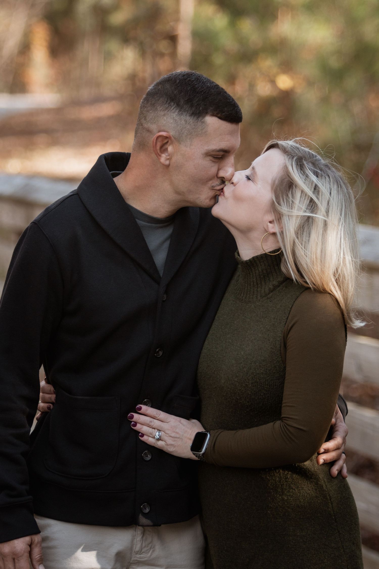 Couple sharing a kiss on a sunlit wooden boardwalk surrounded by autumn trees.