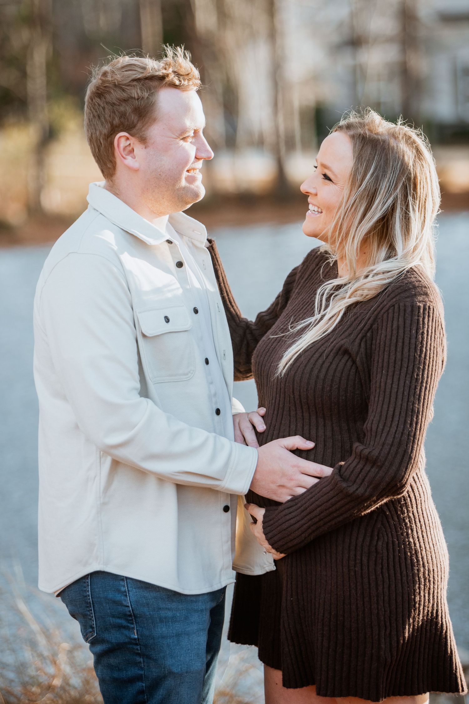 Happy expecting couple embraces and smiles at each other near a peaceful lakeside setting.