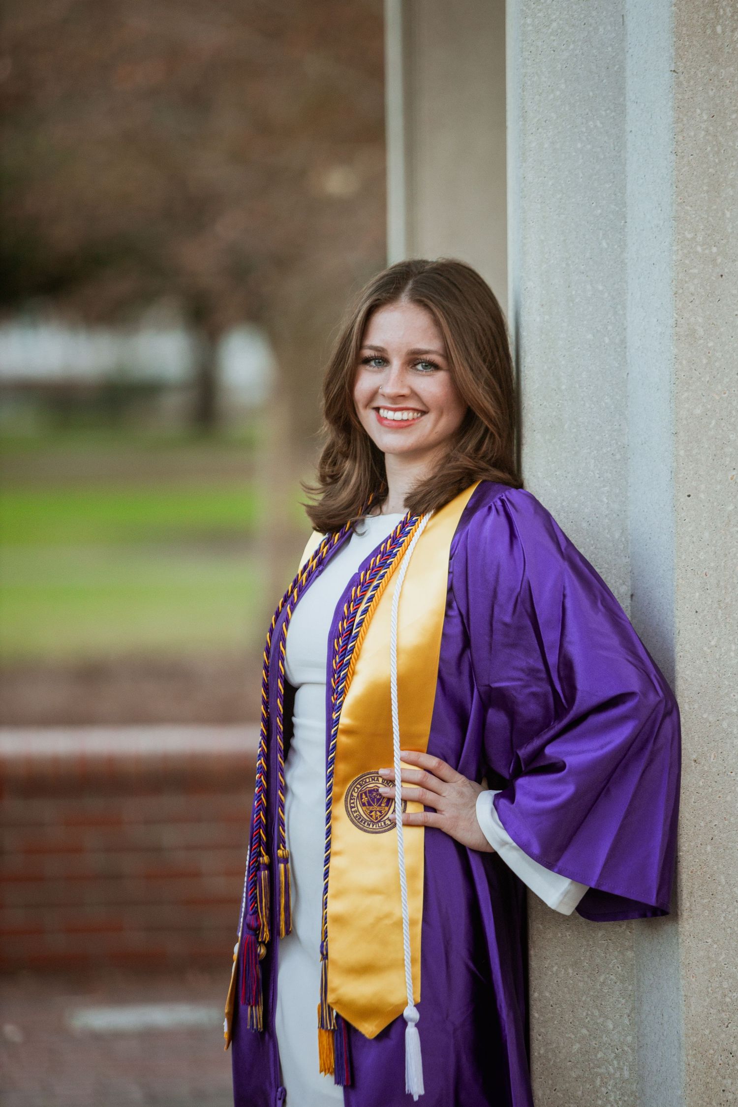 Smiling ECU graduate in purple robe with gold stole leans against white column on campus.