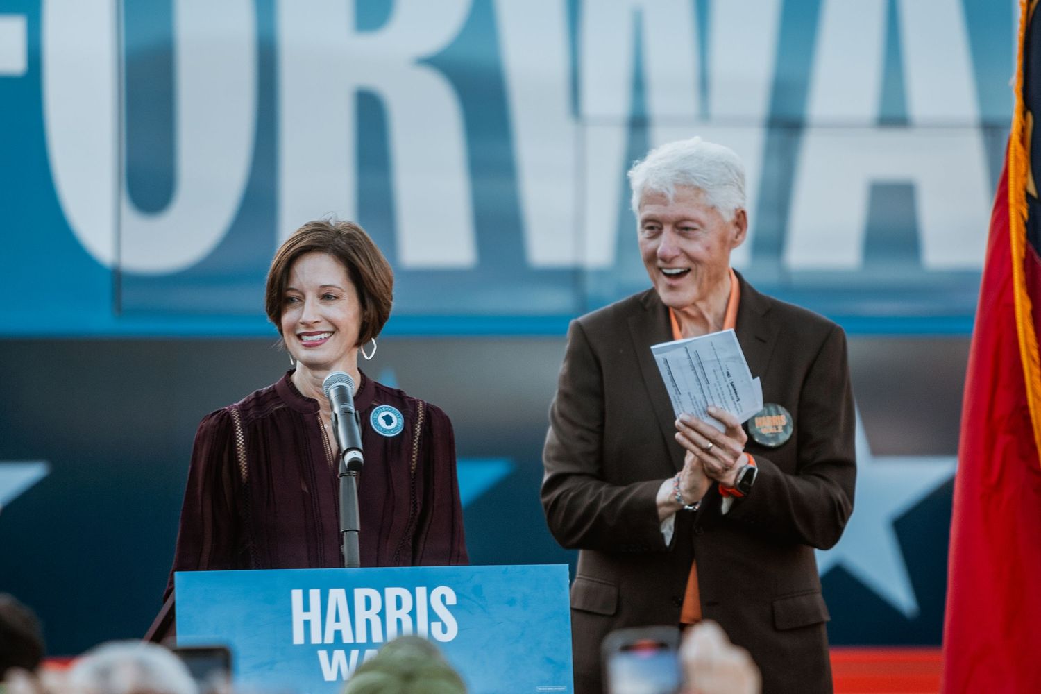 Two people smile together at a Harris-Walz Forward campaign event at an outdoor political rally.