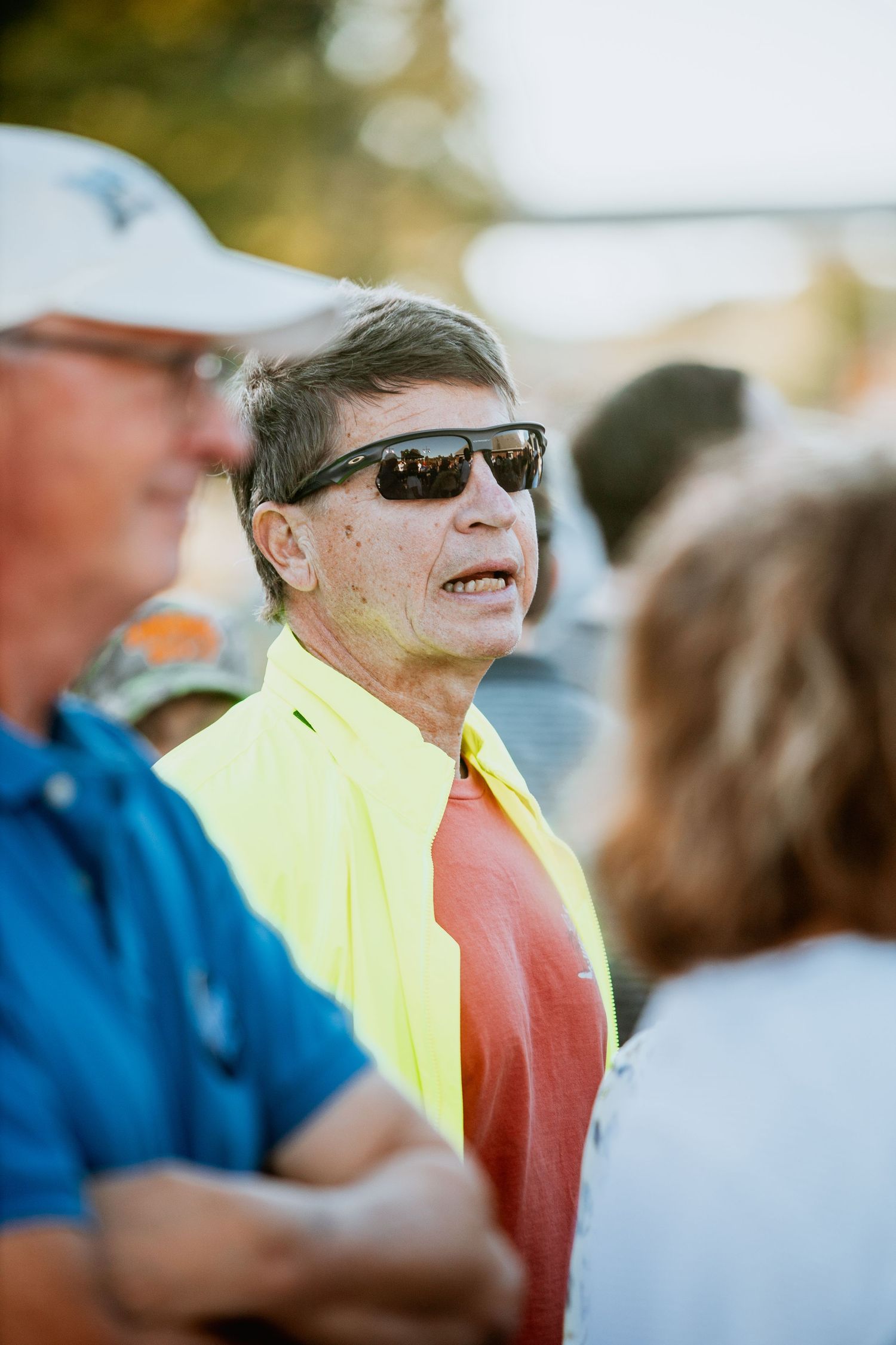 Man in yellow shirt and sunglasses speaking outdoors in a crowd.