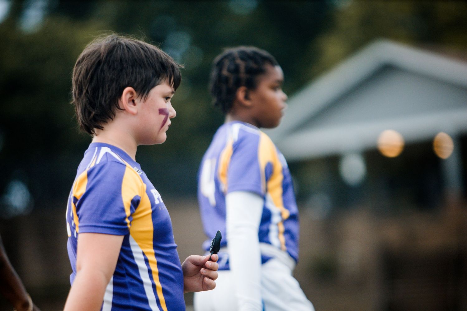 Two young athletes in purple and gold jerseys stand on the sideline during a youth football game.