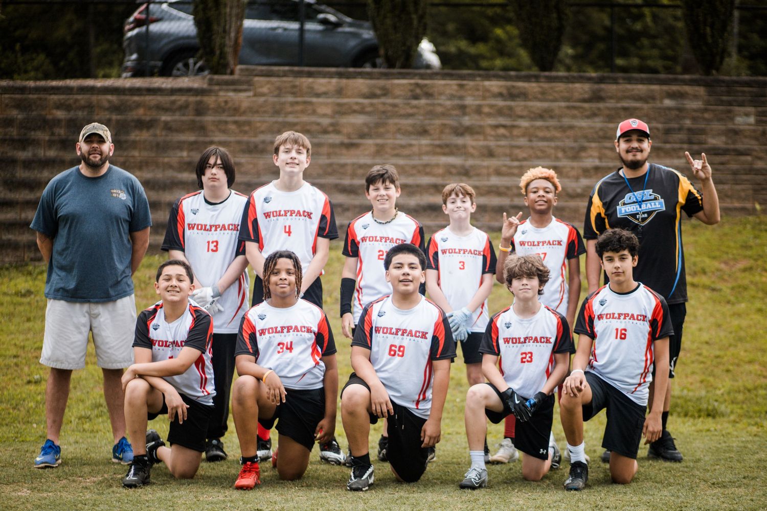 Youth flag football team in red and white Wolfpack jerseys posing with coaches outdoors.