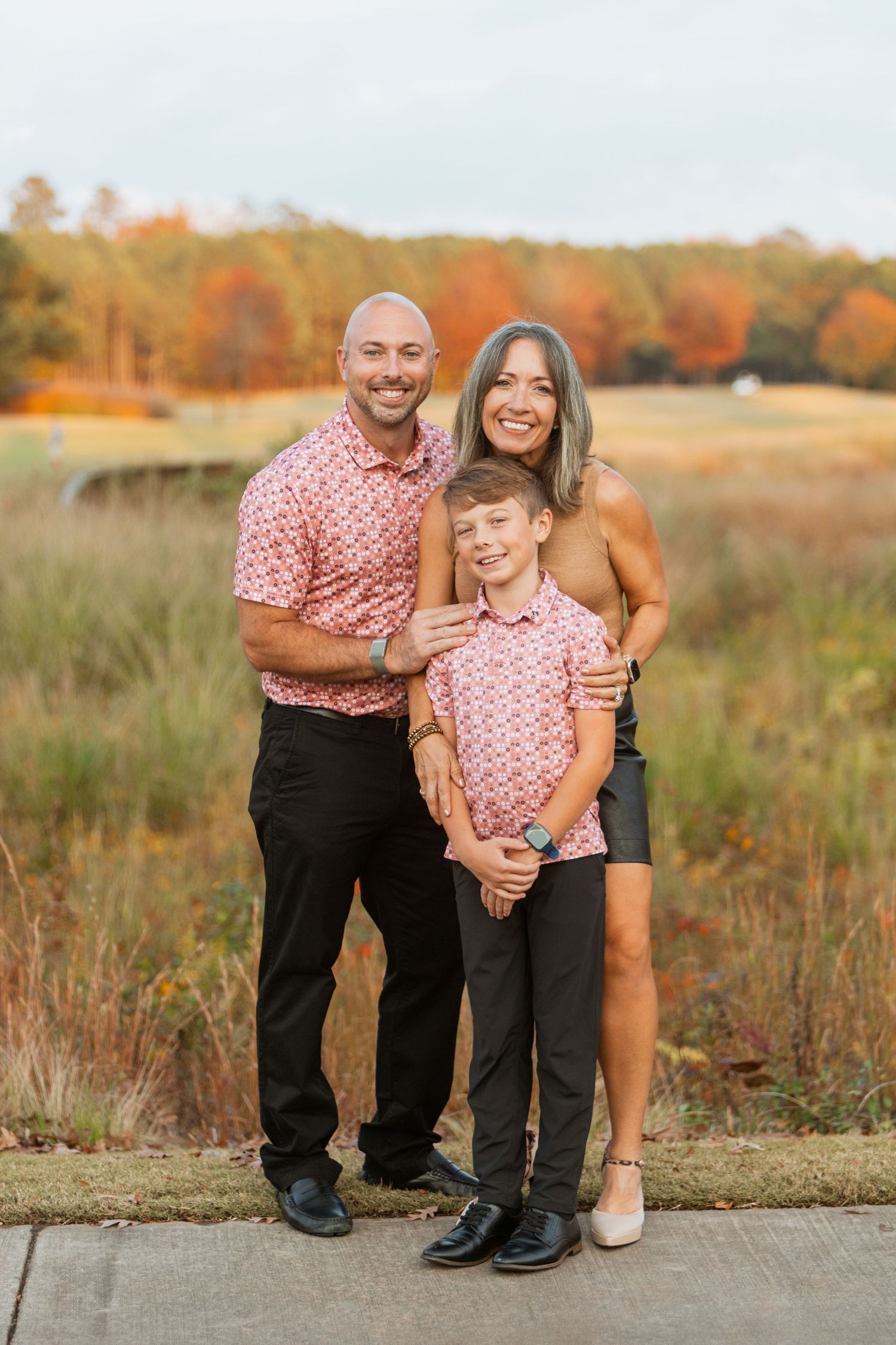 Family of three poses outdoors in matching pink floral shirts during autumn fall photo session.