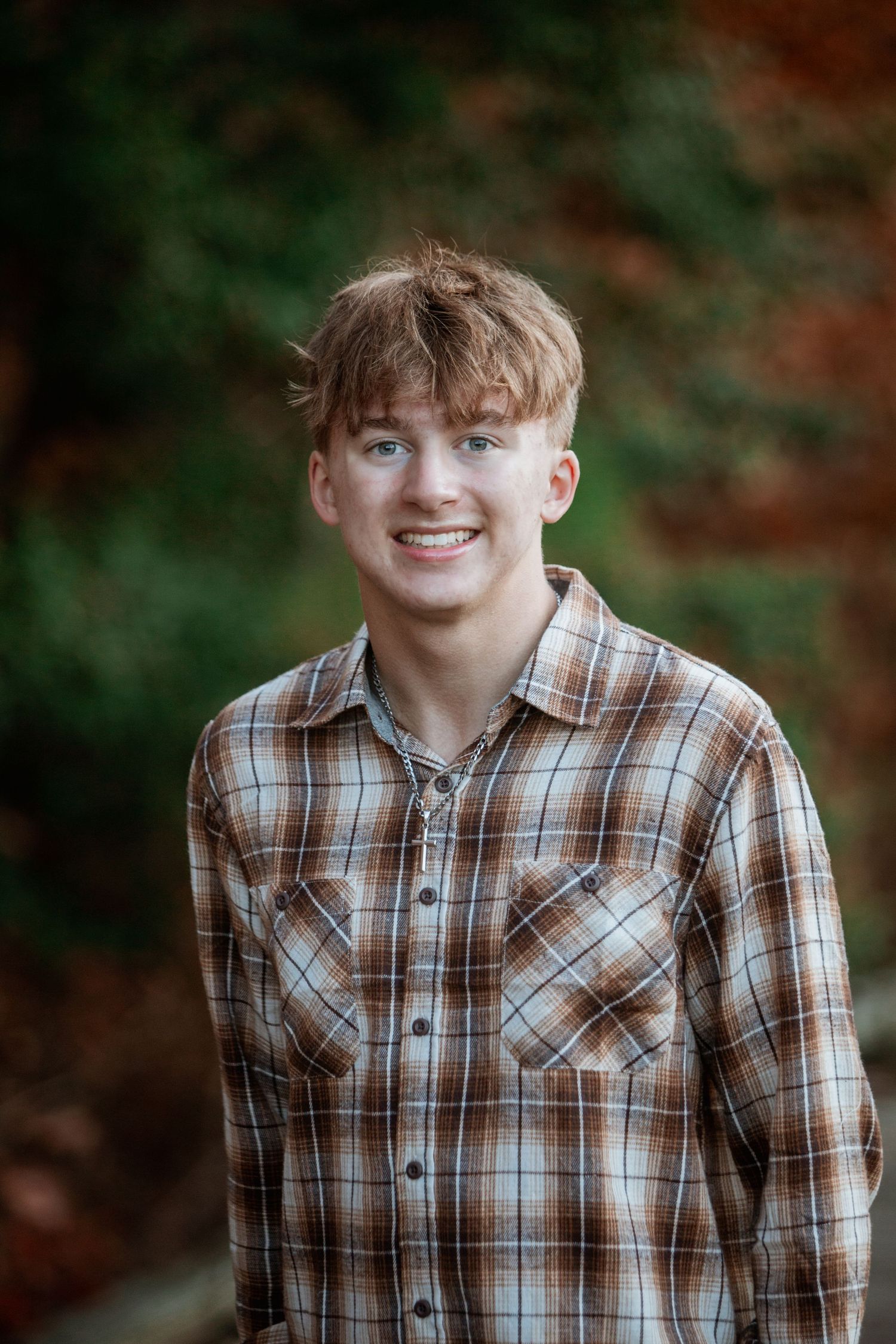 Smiling teenage boy in brown plaid shirt posing outdoors with colorful autumn foliage behind him.