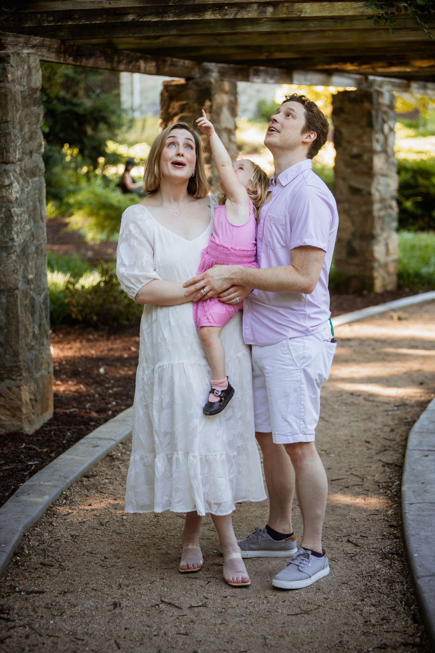 Family of three looking up with curiosity as toddler points skyward under a rustic stone pergola.