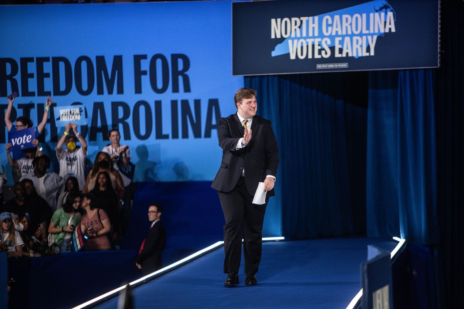 A speaker addresses a crowd at a North Carolina Votes Early political rally on a blue-lit stage.