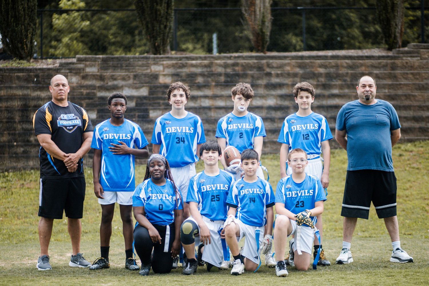 Youth flag football team in blue Devils uniforms posing with two coaches on a grass field.