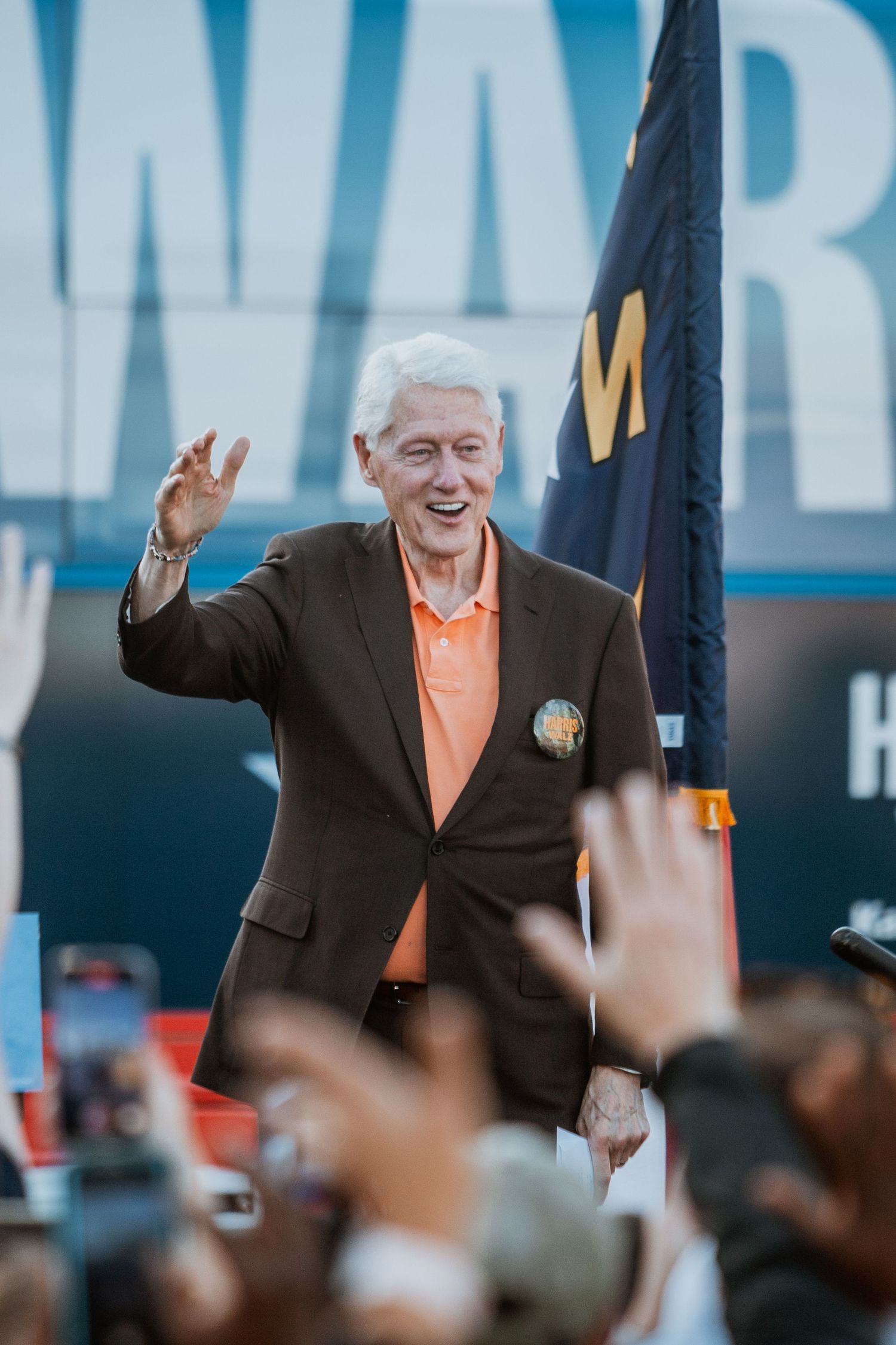 Smiling elderly man in brown blazer waving to crowd at outdoor event.