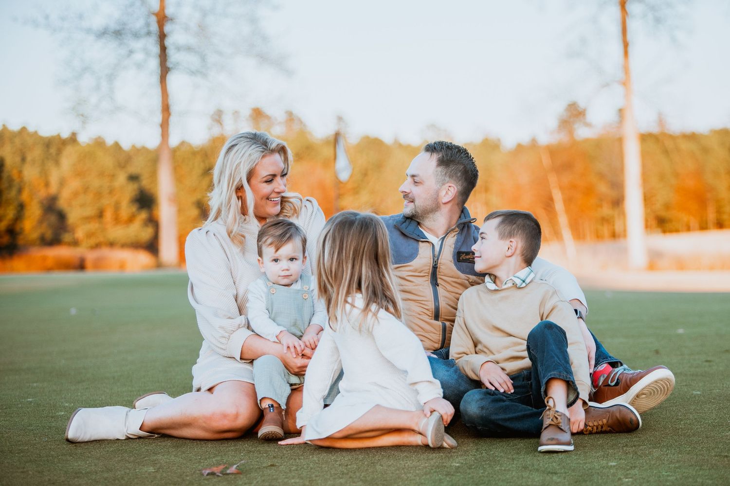 Family of five sitting together on grass in warm autumn sunlight outdoors.