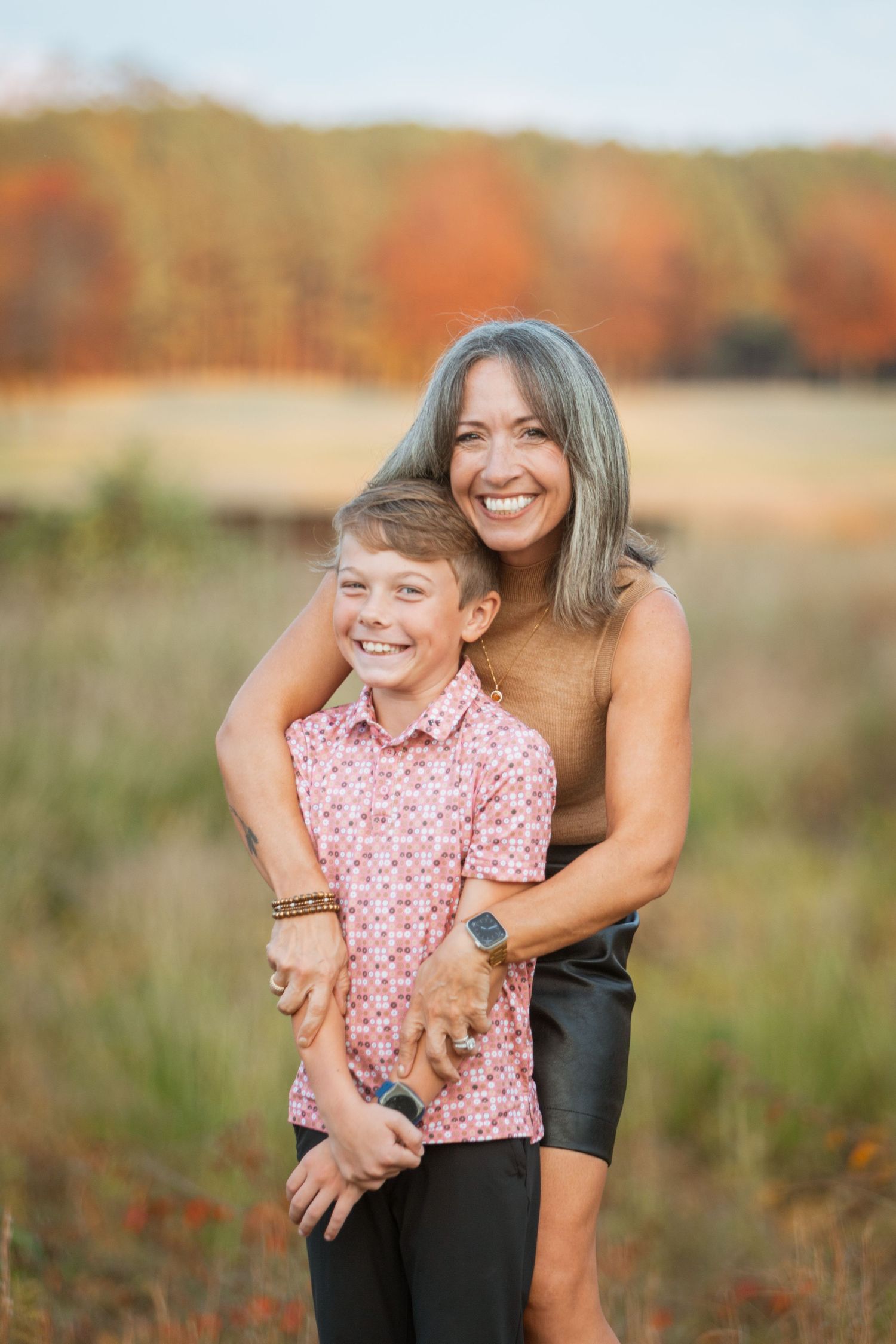 Mother with gray hair hugs smiling son in pink shirt during fall outdoor photo session.
