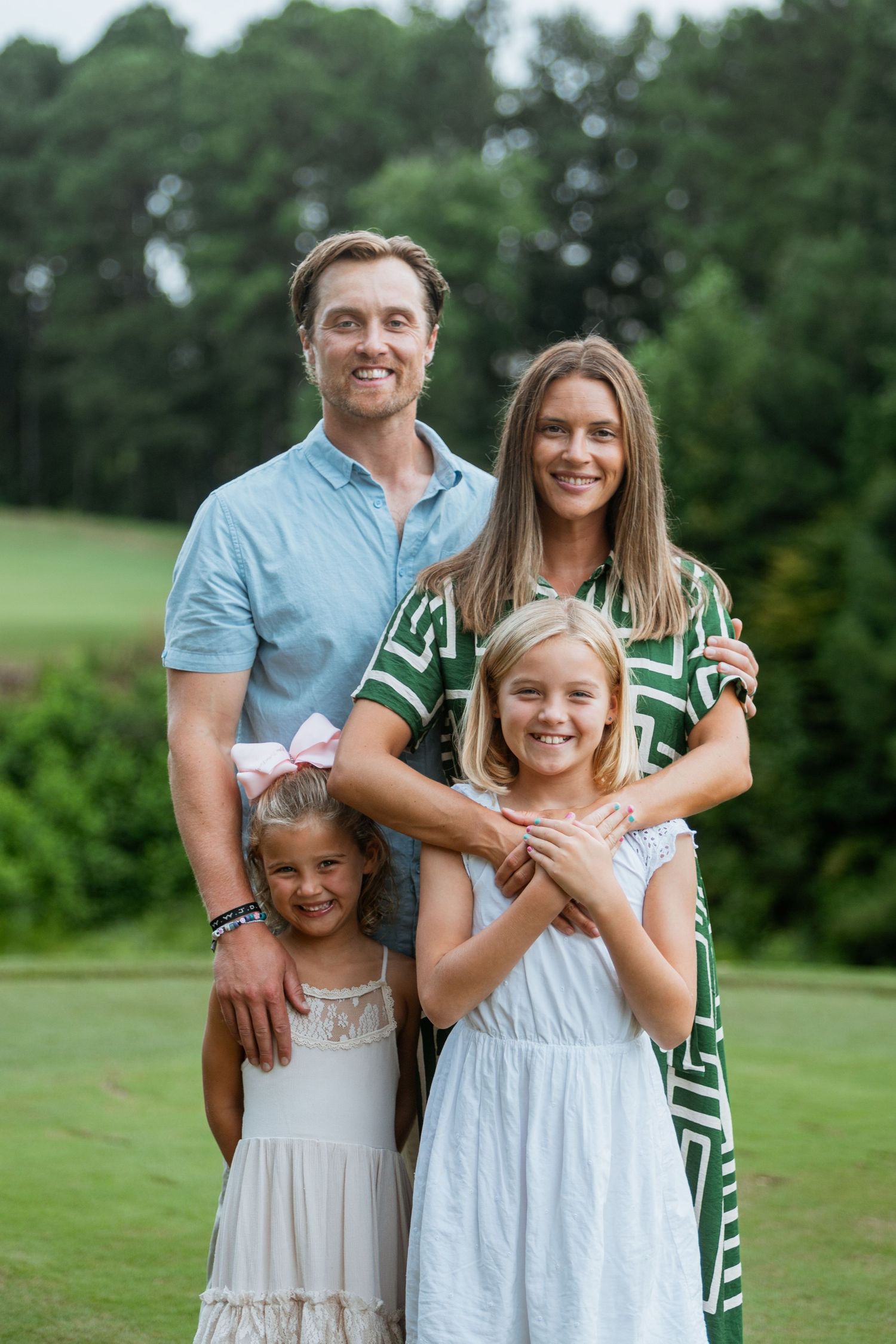 Smiling family of four with two blonde daughters poses together on lush green golf course lawn in summer.