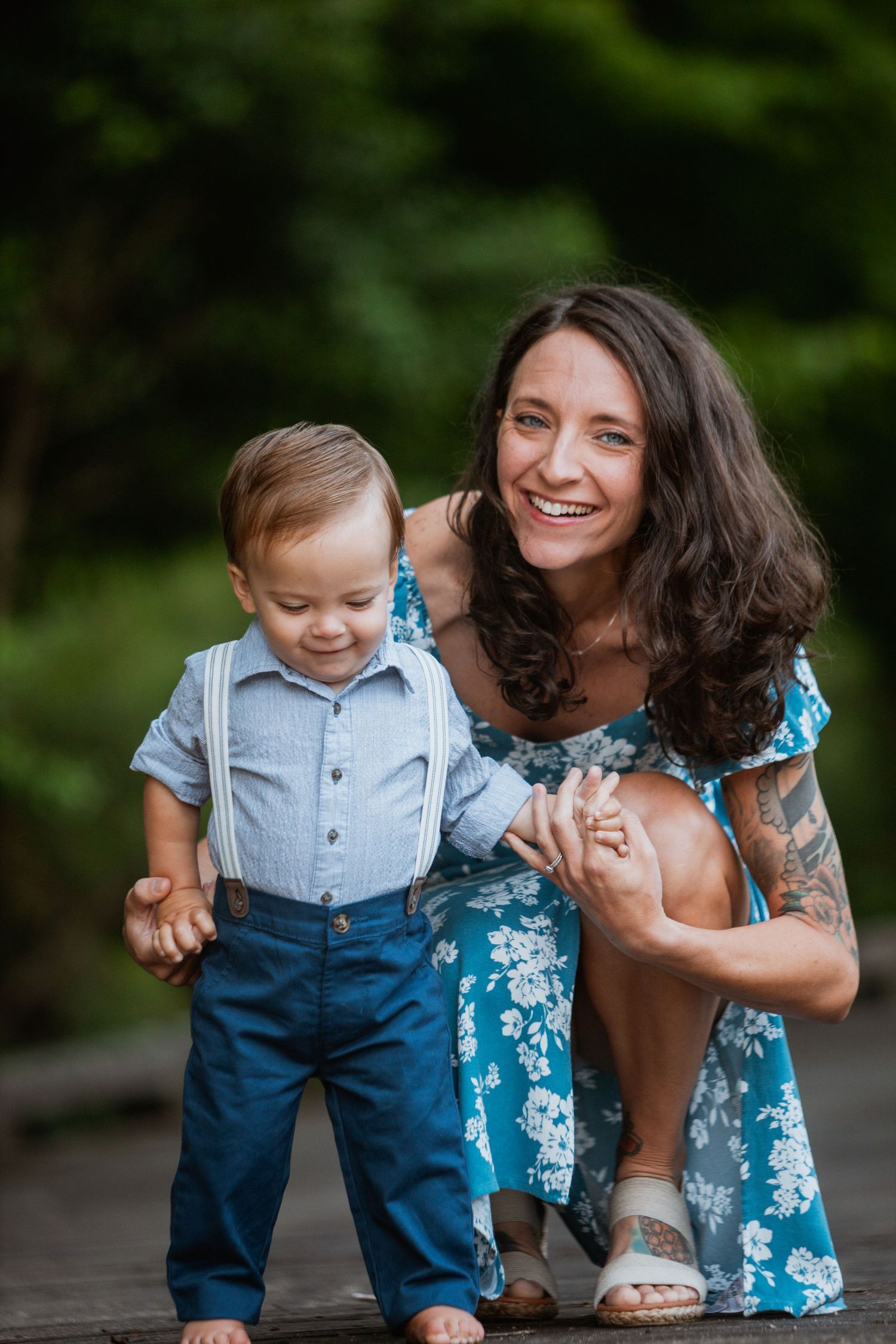 Laughing mother in blue floral dress kneeling beside her toddler son wearing suspenders outdoors.