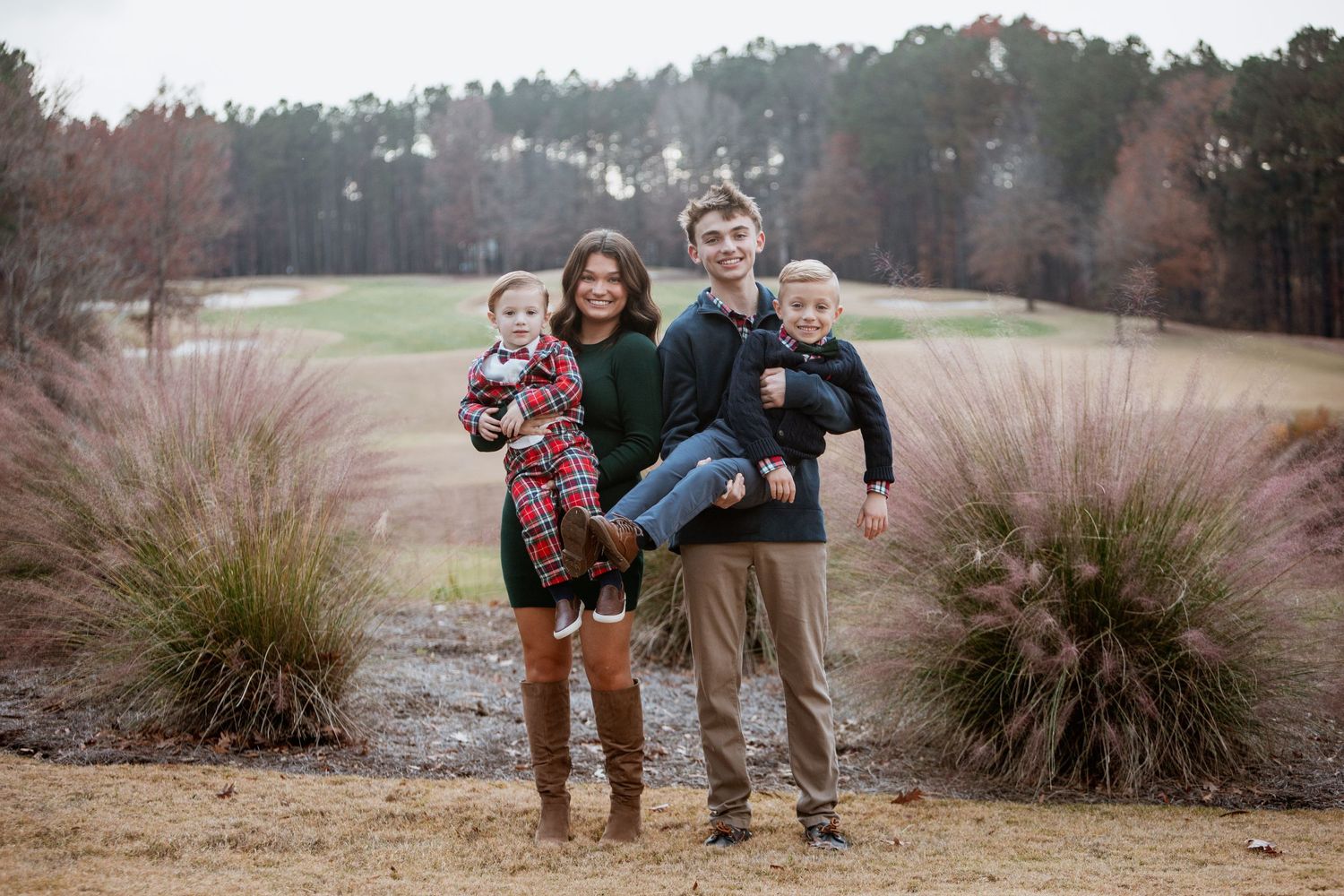 Family of four poses outdoors in holiday attire, two teens each holding a young child near ornamental grasses.