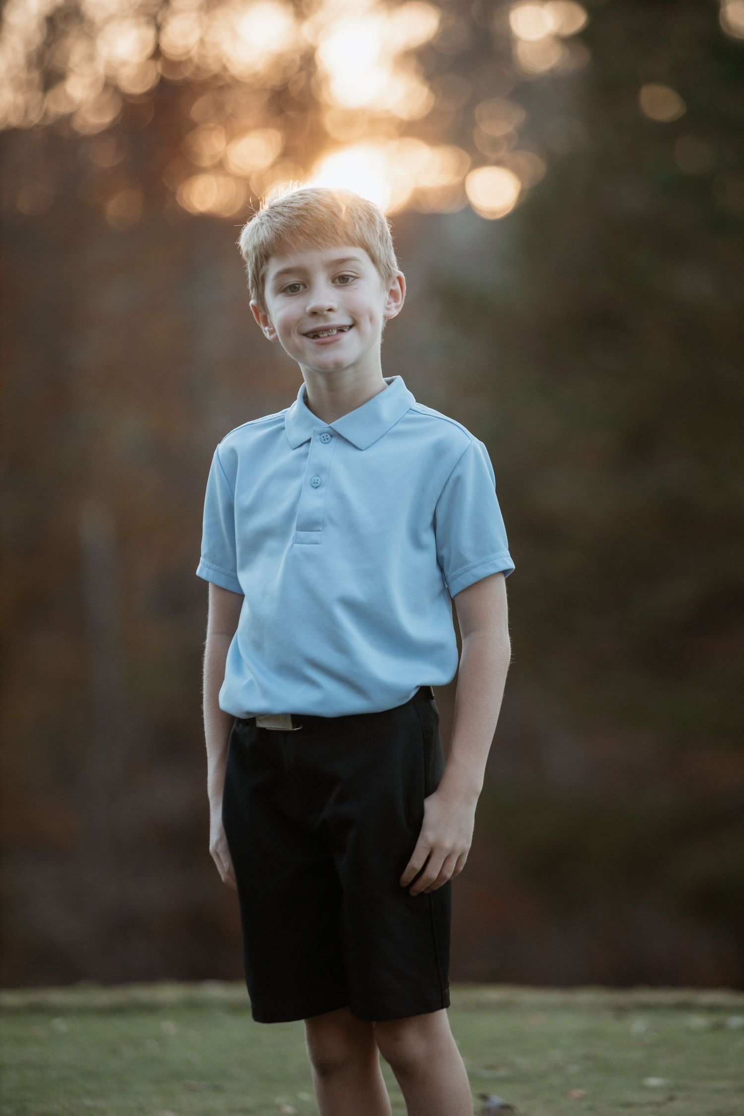 Young blonde boy in a light blue polo and black shorts smiling outdoors at golden hour.