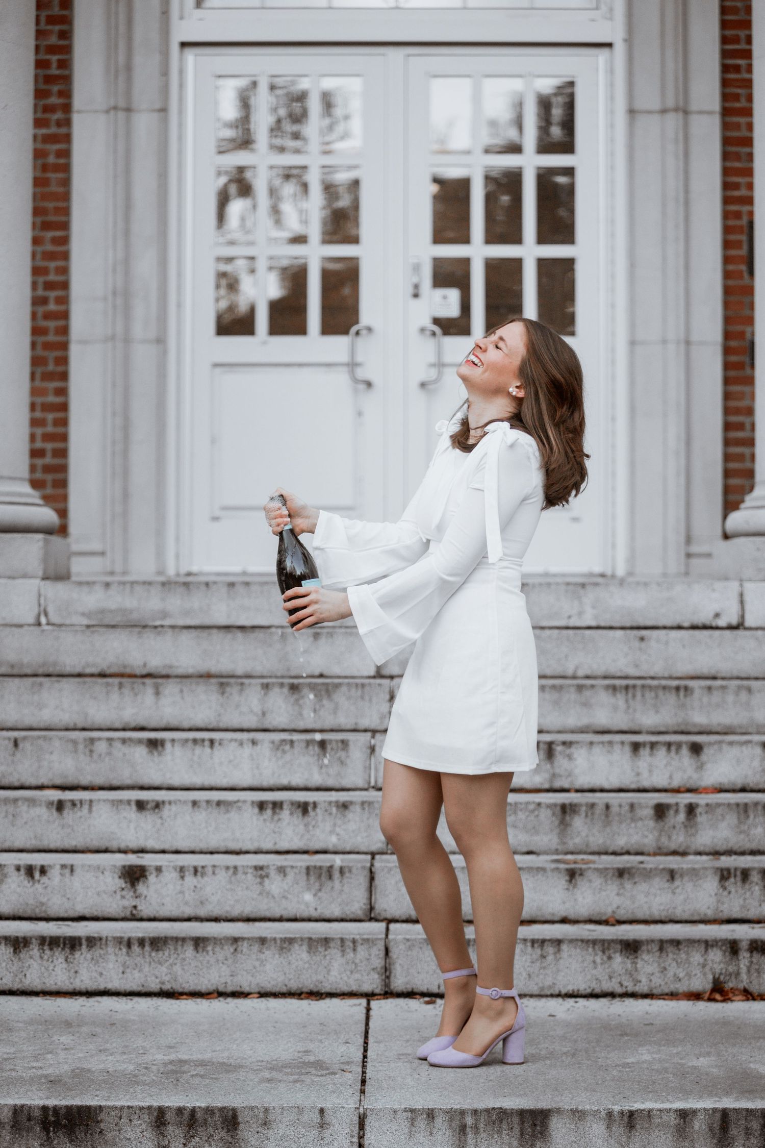 Young woman in white dress celebrates graduation by popping champagne bottle on brick building steps.