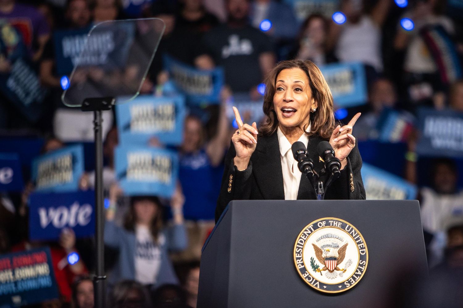 A female politician gestures with both index fingers raised while speaking at a Harris-Walz campaign rally.