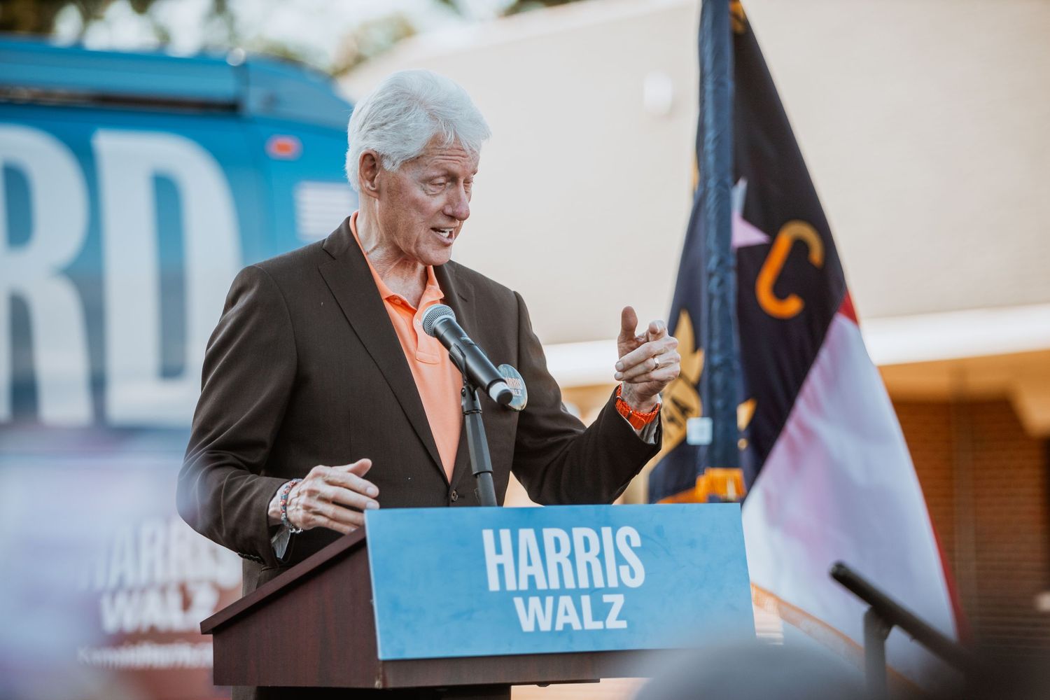 Older man in orange shirt speaks at Harris Walz campaign podium outdoors.