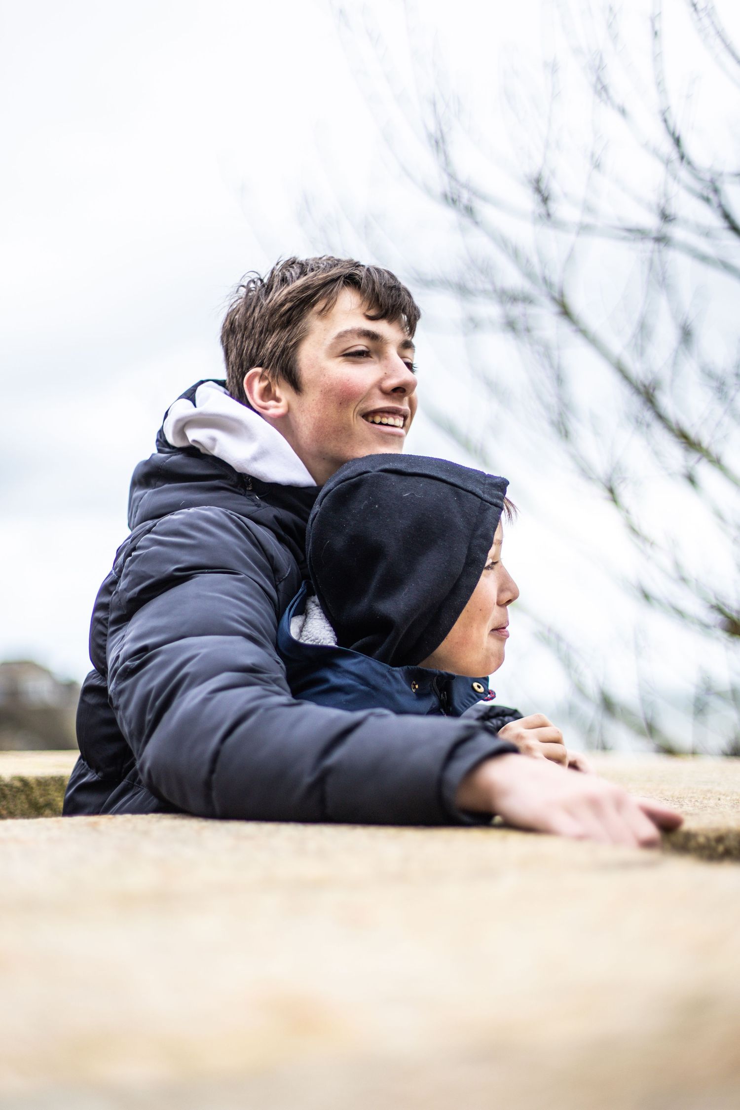 Two smiling boys in navy jackets leaning on a stone wall outdoors with bare trees in the background.