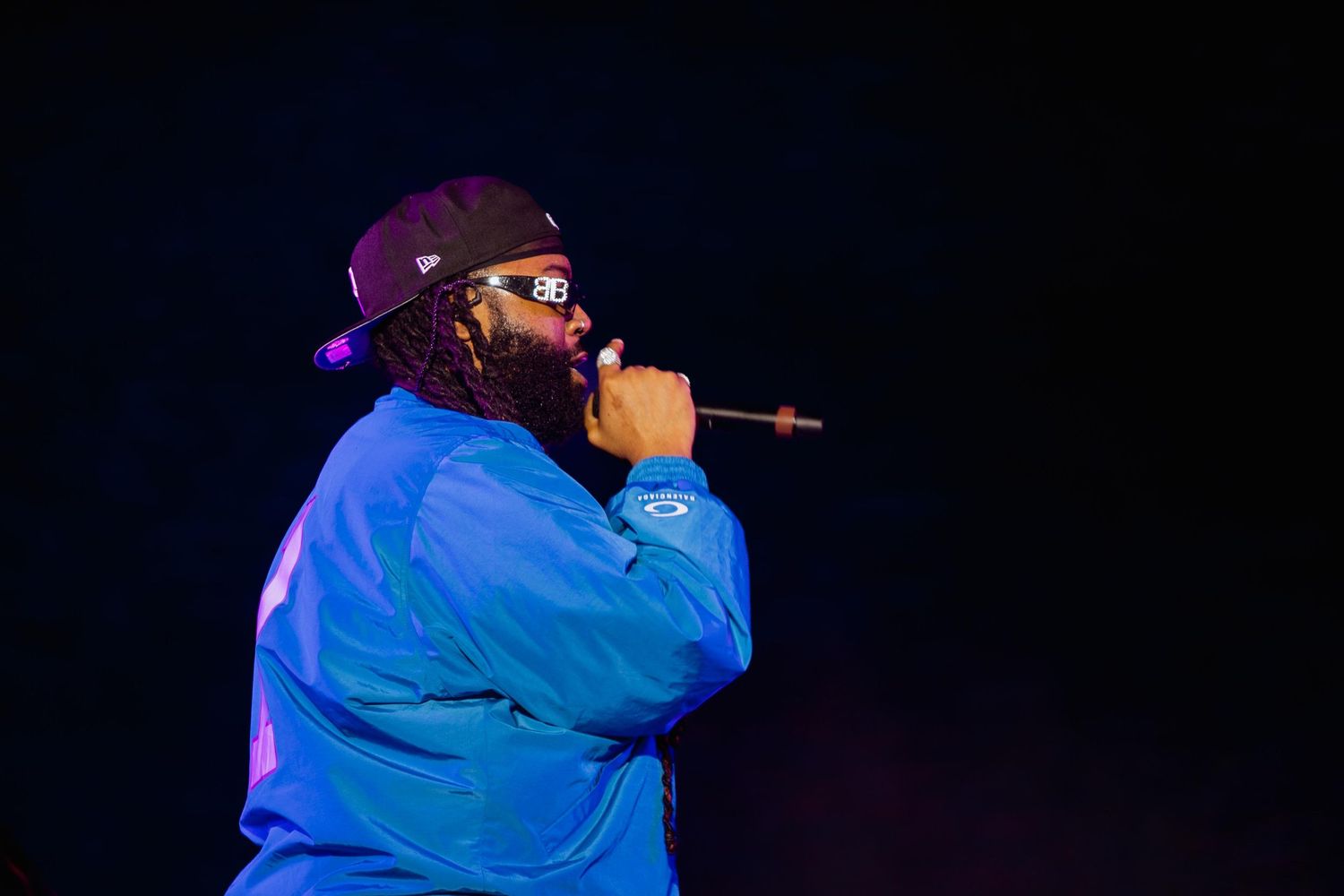 Rapper in blue jacket and cap performing on dark stage.