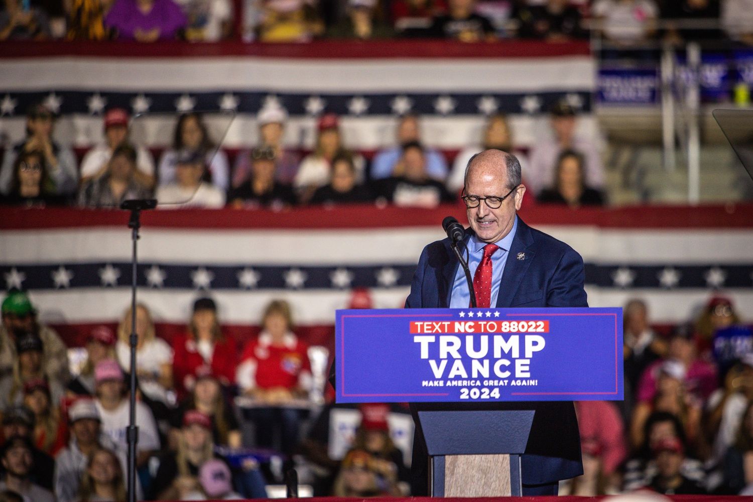 Man speaks at Trump Vance 2024 campaign rally podium before large audience.