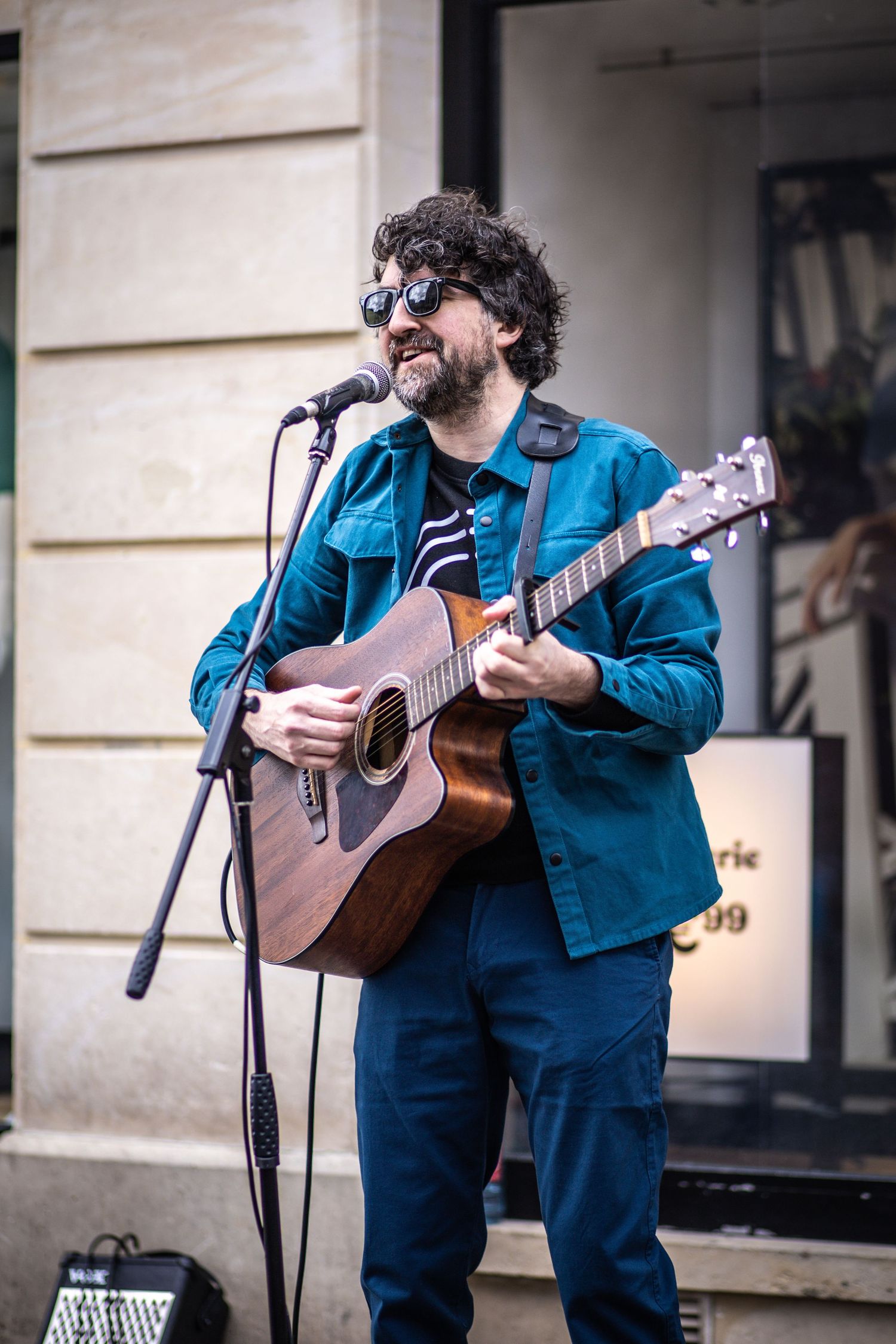 Bearded male street musician wearing sunglasses playing acoustic guitar and singing into a microphone outdoors.