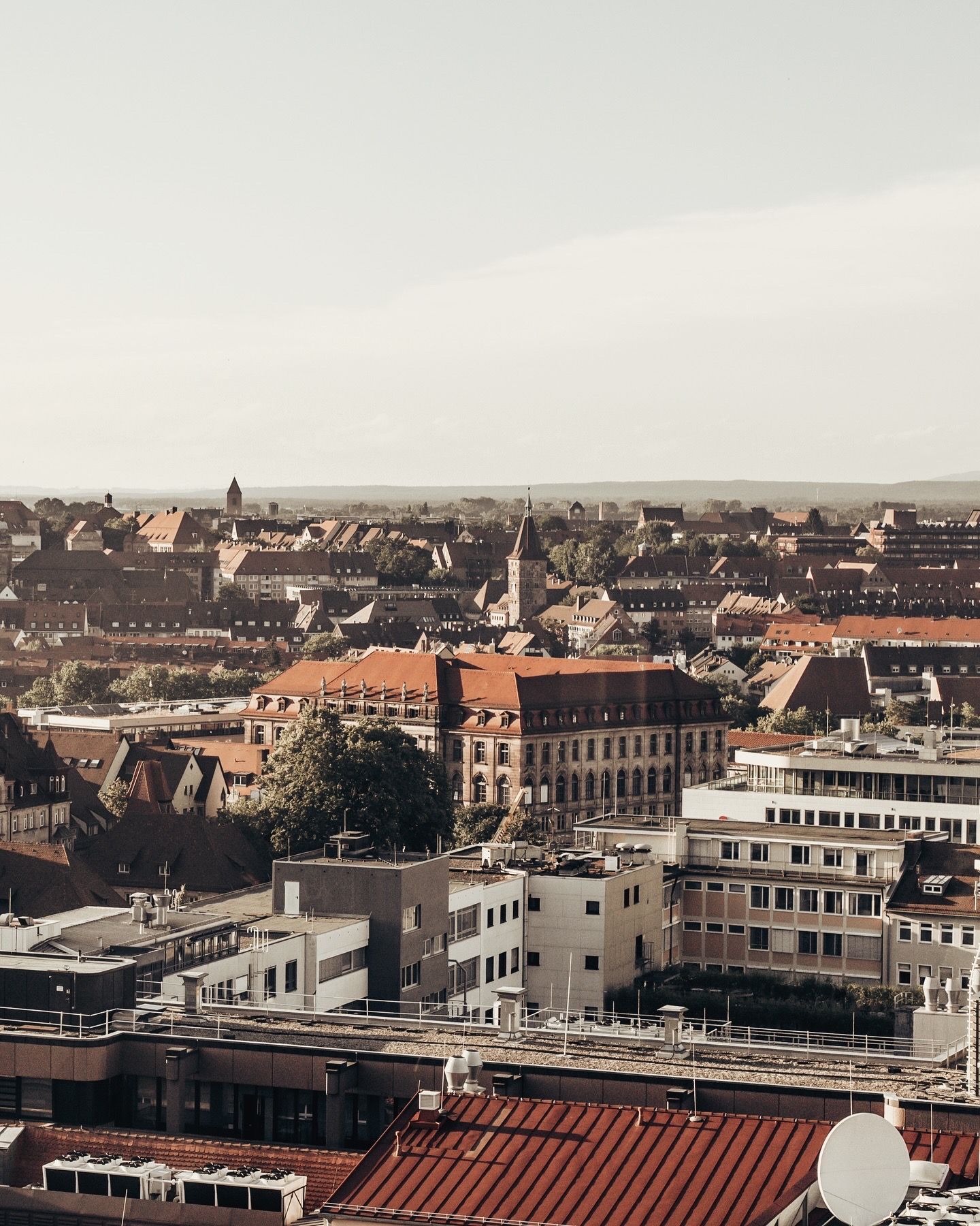 Aerial view of a European city skyline with red-roofed buildings and a church spire under a hazy sky.