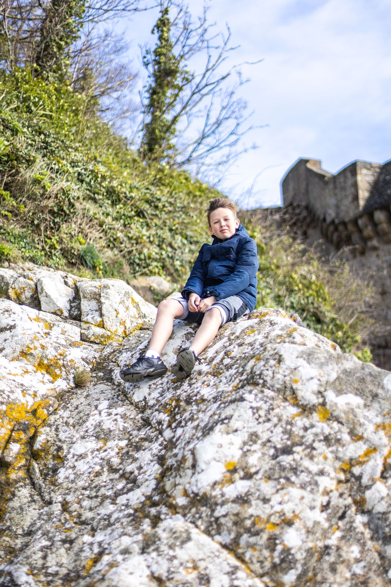 Young boy sitting on mossy rocks near ancient stone walls surrounded by lush green vegetation.