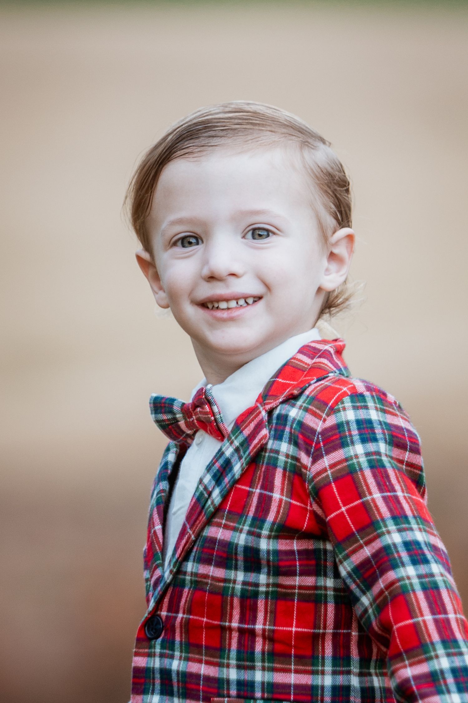 Close-up portrait of smiling young boy in red plaid jacket and bow tie outdoors.