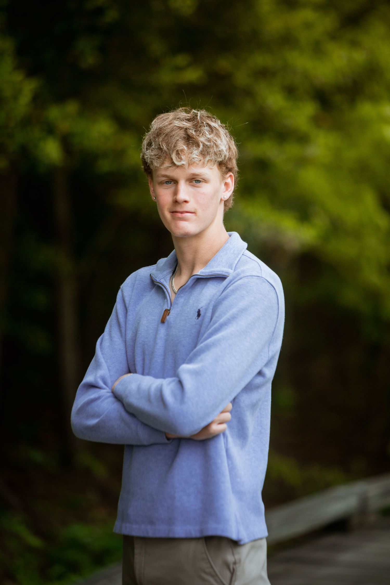 Young man with curly blonde hair stands arms crossed wearing light blue pullover on wooded path.