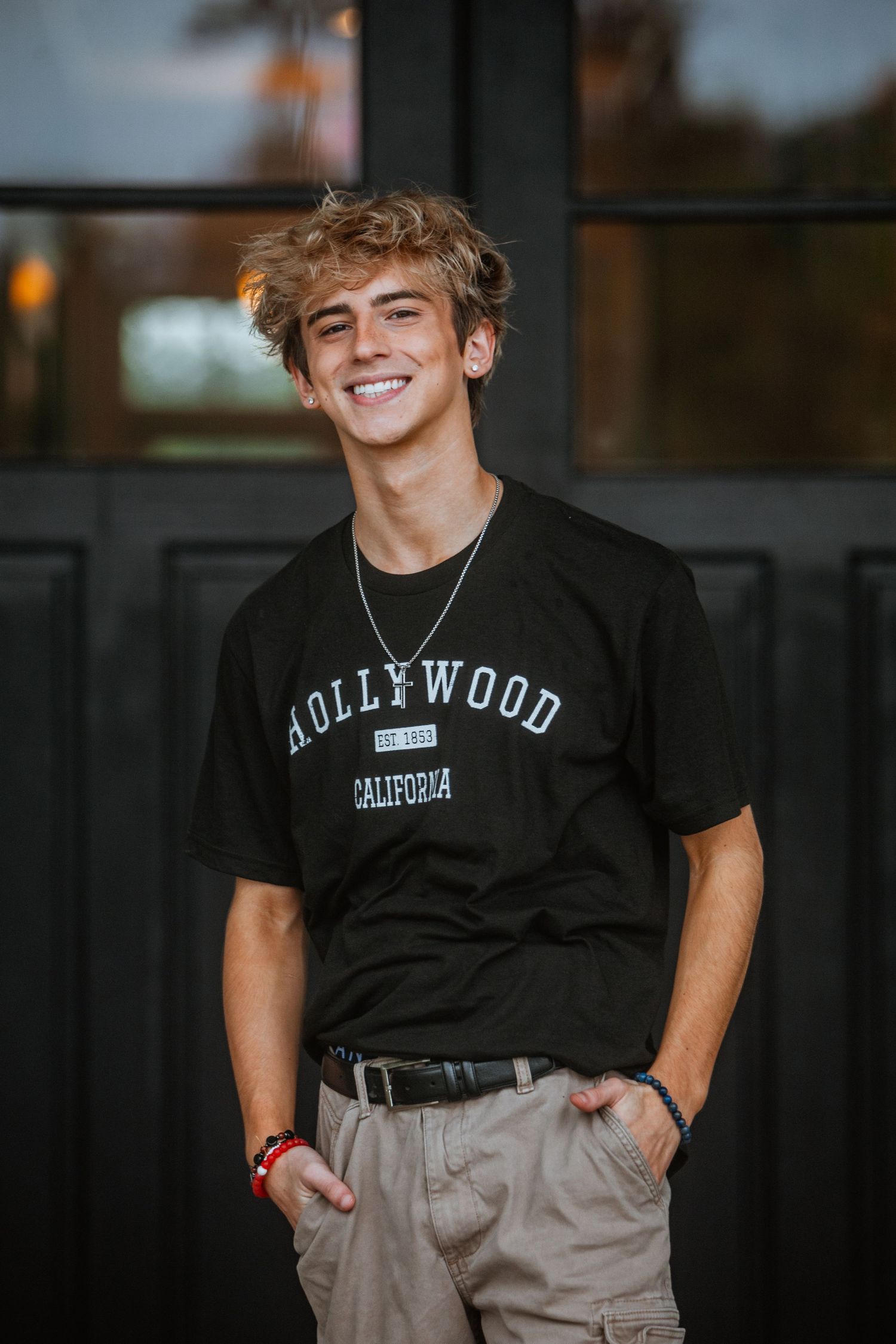 Smiling teenage boy with blonde hair in black Hollywood t-shirt standing outdoors.