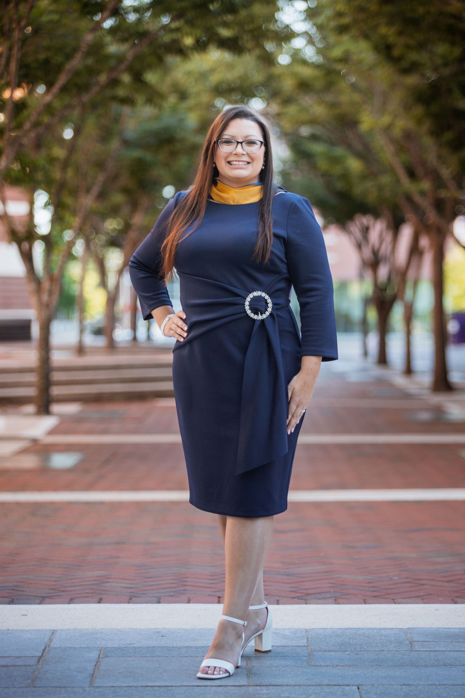 Professional woman in navy blue dress with gold turtleneck posing outdoors on brick walkway.
