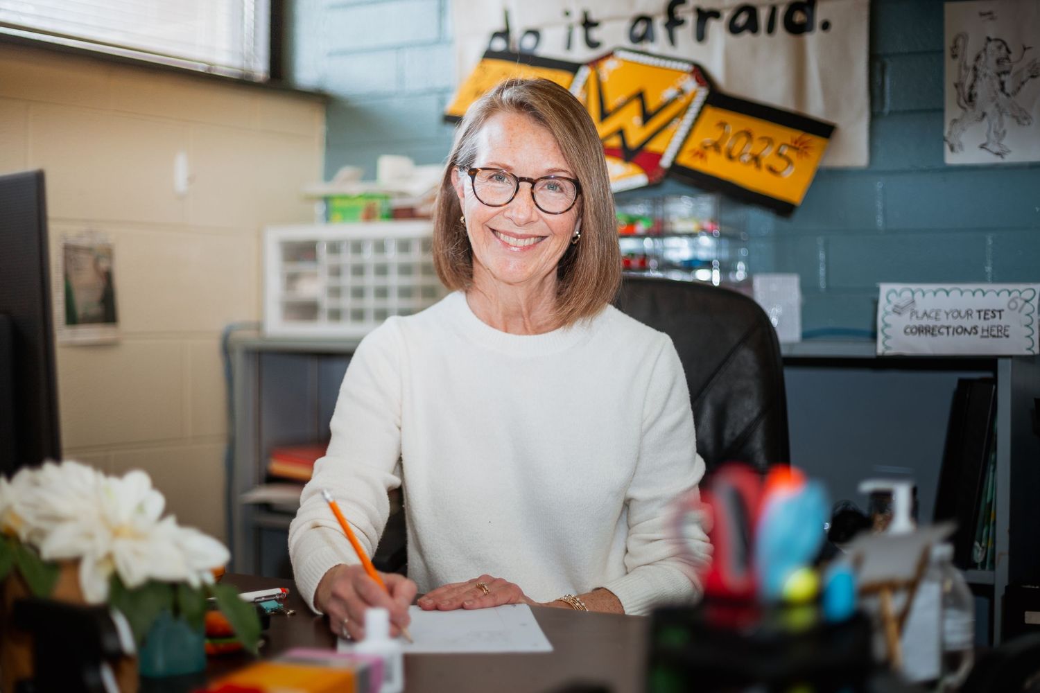 Smiling older woman with glasses sitting at a desk in a classroom office setting.
