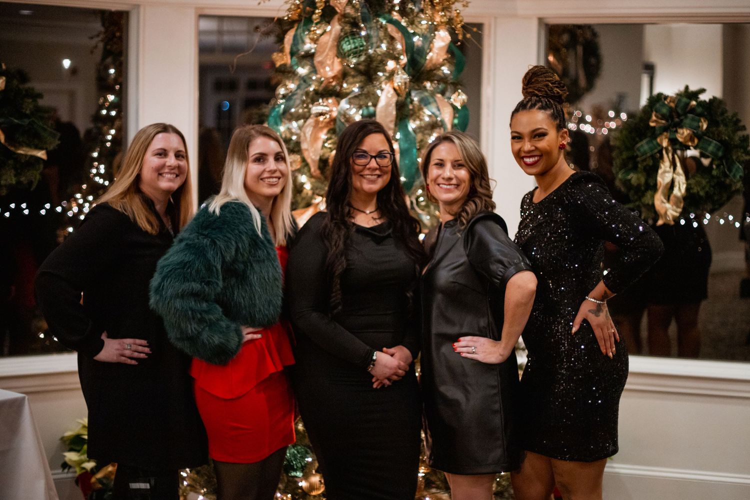 Five women posing together in festive outfits in front of decorated Christmas tree at holiday party.
