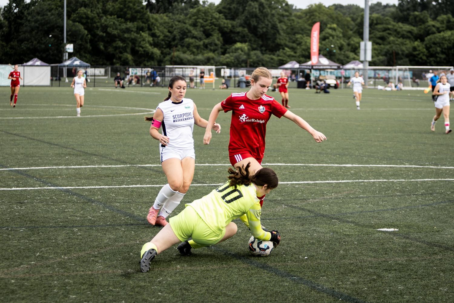 Goalkeeper in green dives to make save as red and white team players compete in womens youth soccer match.